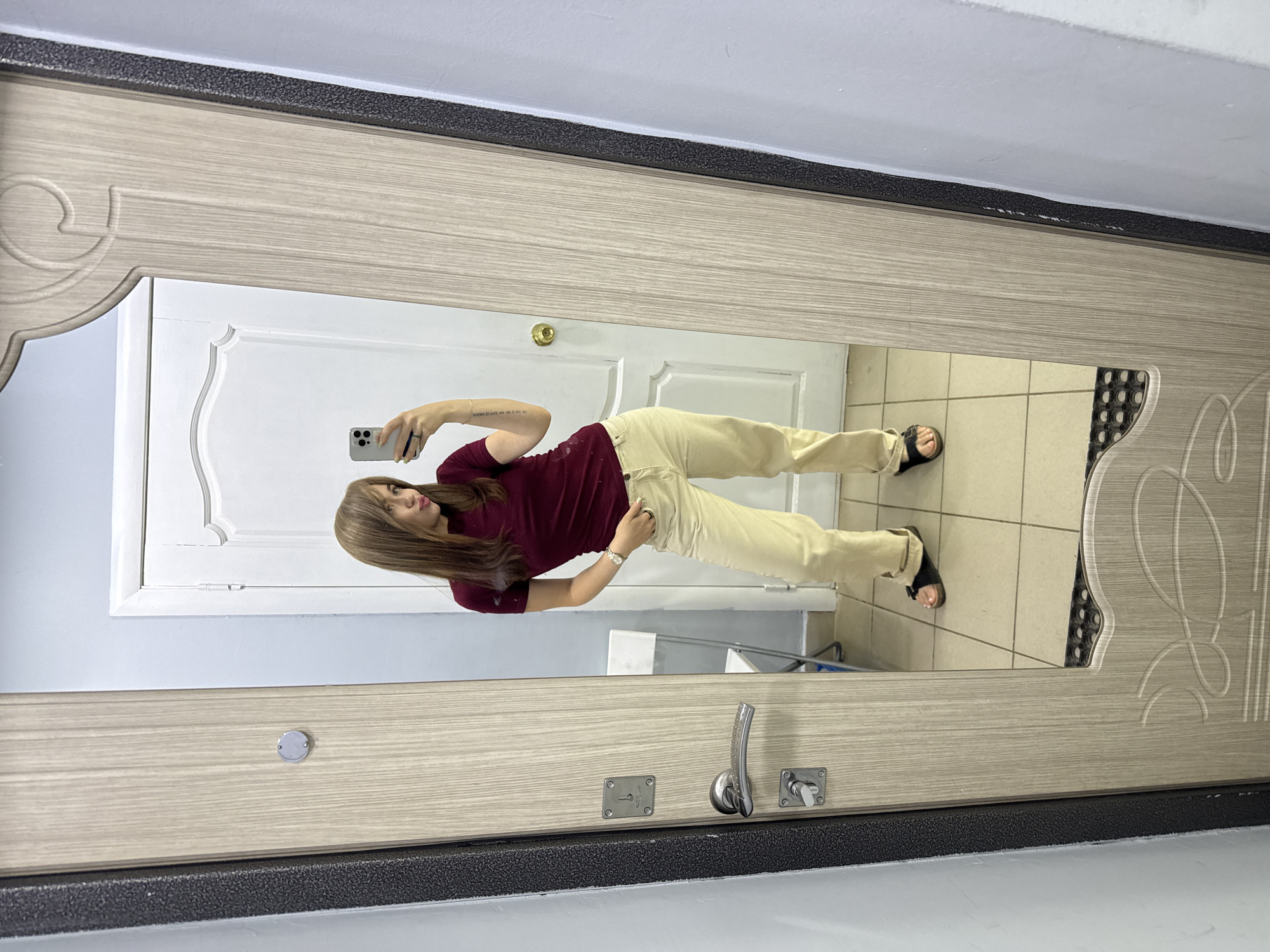A young woman with medium-length brown hair is taking a mirror selfie in what appears to be a doorway. She is wearing a maroon short-sleeved t-shirt and light beige pants, along with black sandals. Her right hand is in her pocket, and she is holding her phone in her left hand. The mirror reflects a white door behind her, as well as a small white chair. The doorframe is made of light-colored wood with decorative carving at the top. To the left of the mirror is a dark grey door edge with a textured surface. The floor is tiled with light-colored square tiles. The lighting is neutral, suggesting an indoor environment. There is no information in the image to suggest a specific location in Chaikovsky, Russia.