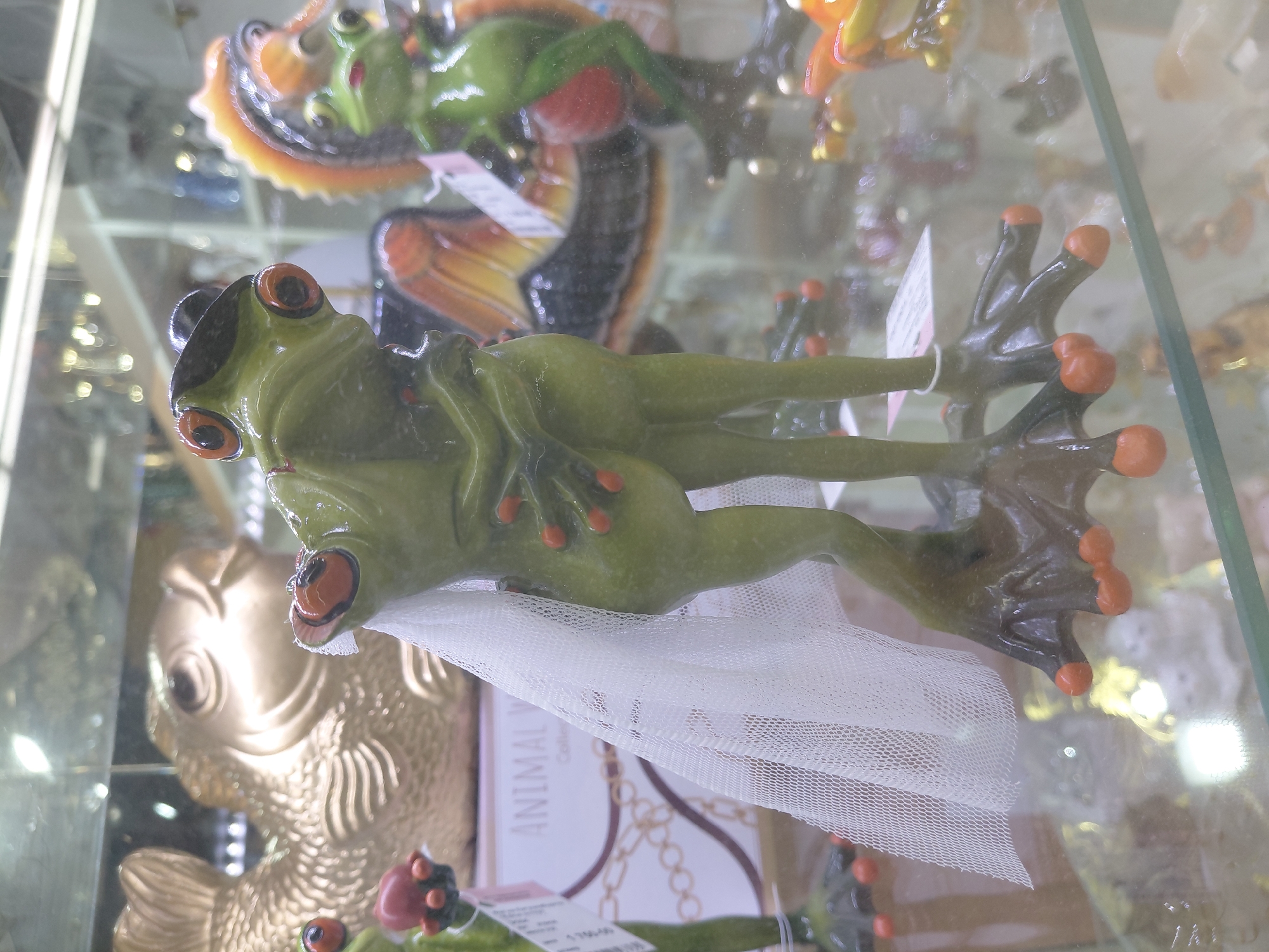 The image displays a close-up view of a glass display case containing various ceramic figurines. The primary focus is on a pair of green ceramic frogs, posed as if embracing. The frog on the left is wearing a black hat and a white veil, suggesting a wedding theme. The frog on the right is partially obscured by the first frog, but appears to be a matching figurine. Both frogs have red accents on their fingers and toes.
Behind the frogs, a large, golden ceramic fish figurine is visible. To the right of the frogs, another green ceramic frog figurine with an orange crest is positioned. Several other blurred figurines and decorative elements are present in the background, also behind the glass. The lighting in the display case creates reflections and highlights on the glass and figurines. The overall scene appears to be in a retail environment, possibly a gift shop or a collector's store. The figurines are placed on a glass shelf within the display case.