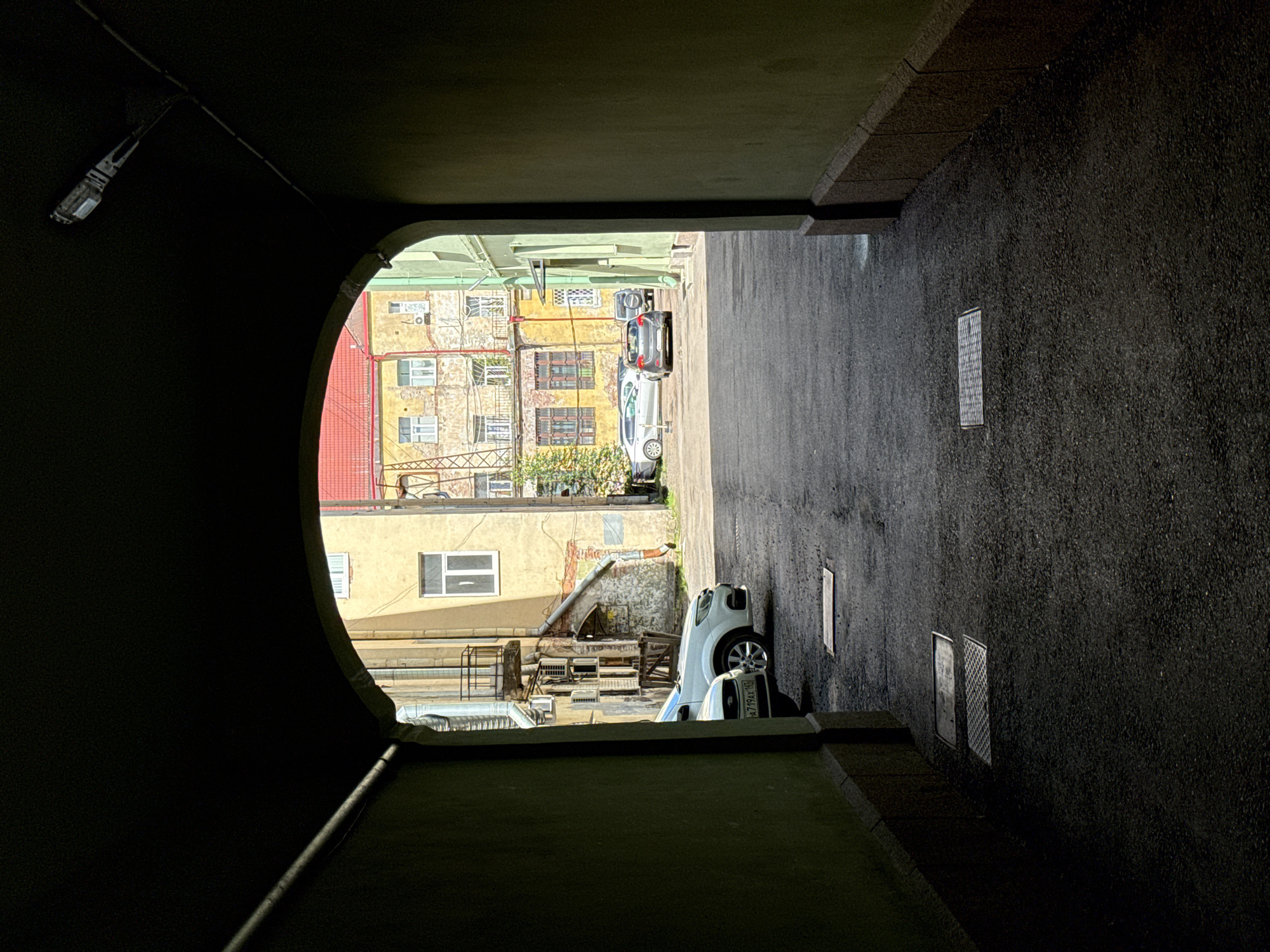The image shows a view through a dark, arched passageway, opening onto a sunlit courtyard in Vyborg, Russia. The passageway walls are painted a muted green, with a rough, textured finish at the base. A light fixture is visible on the ceiling of the archway on the right.
The courtyard is paved with asphalt and appears to be a shared space for several apartment buildings. Several cars are parked: a white sedan with a license plate ending in "AX 140" is visible on the left, partially obscured by the passageway's shadow. Further into the courtyard, a gray hatchback is parked, and behind it, a silver sedan and a dark SUV are also present.
The buildings in the courtyard are old, with yellow and pale green facades. Architectural details include balconies with ornate railings, numerous windows of varying sizes, and what appear to be air conditioning units mounted on the walls. One building has a red tiled roof. A metal fire escape or industrial structure is visible on the far left building. The overall impression is one of an older, European city courtyard with signs of residential use and modern vehicles. There are no people visible in the scene.