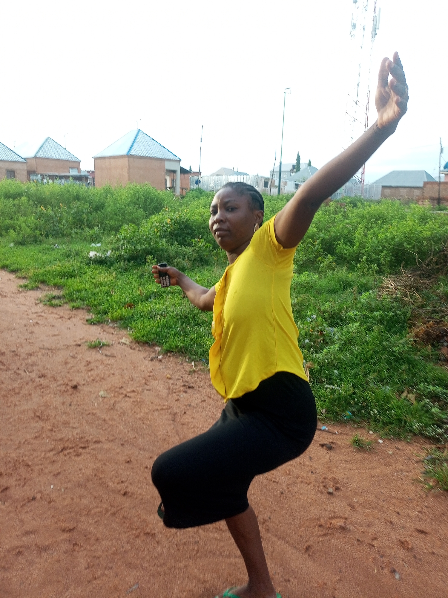 An African woman in a yellow t-shirt and black skirt is standing in a dirt path, with her right arm extended upwards. She is holding a black mobile phone in her left hand. The background shows green vegetation, a few buildings with blue roofs, and a cell tower. The scene is outdoors, under a bright sky.