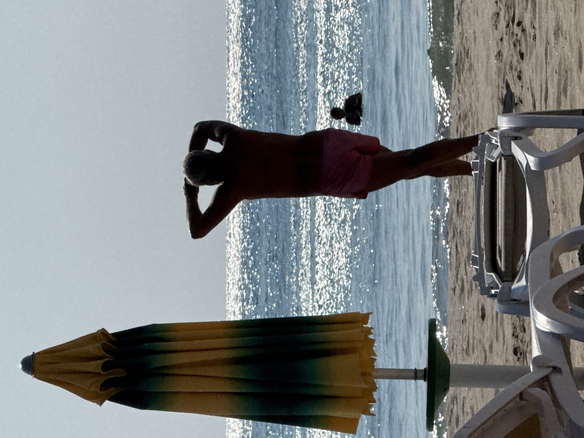In Obrochishte, Bulgaria, a beach scene unfolds under a hazy sky. In the foreground, two white plastic lounge chairs sit on the sand, one partially visible. To the left, a closed beach umbrella, striped yellow and dark green, stands upright on a green base. Beyond the chairs and umbrella, the sea stretches out, its surface shimmering with sunlight. A man, silhouetted against the bright water, stands on the beach with his arms raised behind his head. He is wearing pink shorts. Further out in the water, another person, barely visible as a silhouette, is swimming. The sand is light brown and textured.