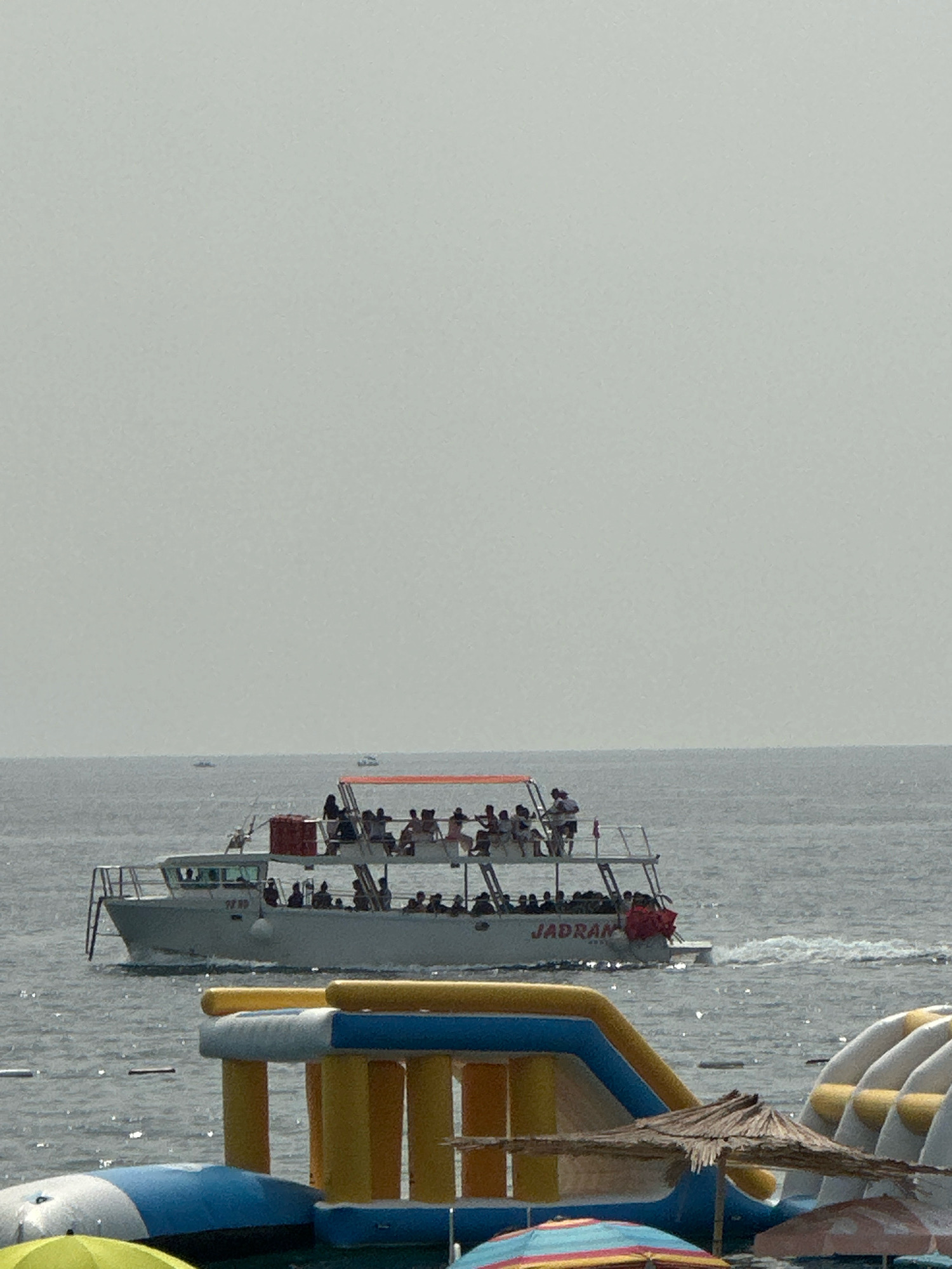 In Boreti, Montenegro, a crowded white passenger boat with an orange awning sails on the sea. The boat, named "JADRAN," carries numerous people on its two decks. In the foreground, inflatable water attractions, including slides and platforms, are partially submerged in the water, suggesting a recreational area. The sky is overcast, and the sea appears calm with small ripples. Several other smaller boats are visible in the distance.