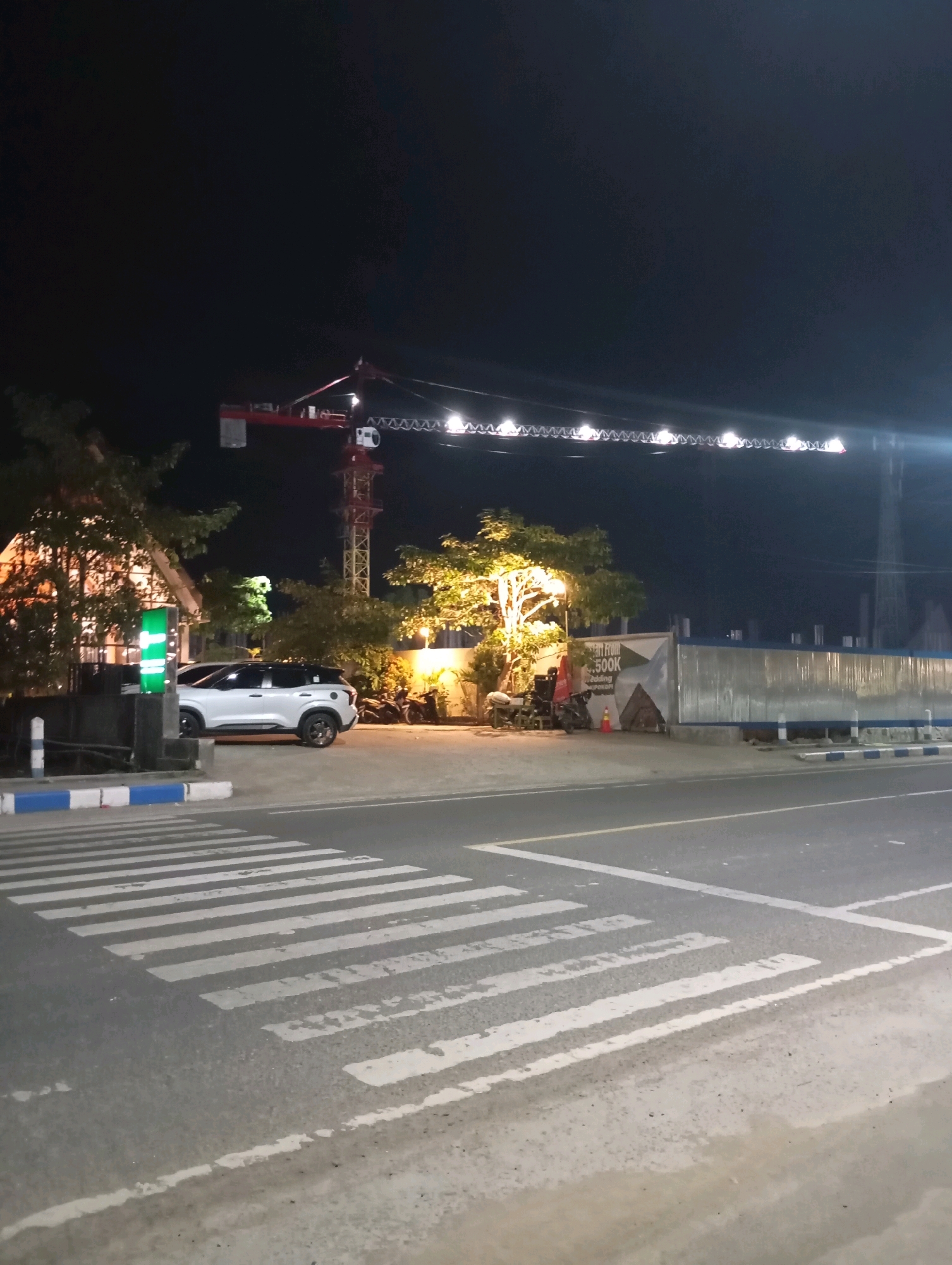 The image shows a nighttime scene in the city of Pacitan, Indonesia, featuring a construction crane with lights illuminating its boom, a white SUV parked near a building with trees and a bright green sign, and a pedestrian crossing in the foreground. The overall environment appears to be a street or road adjacent to a construction site or commercial area.