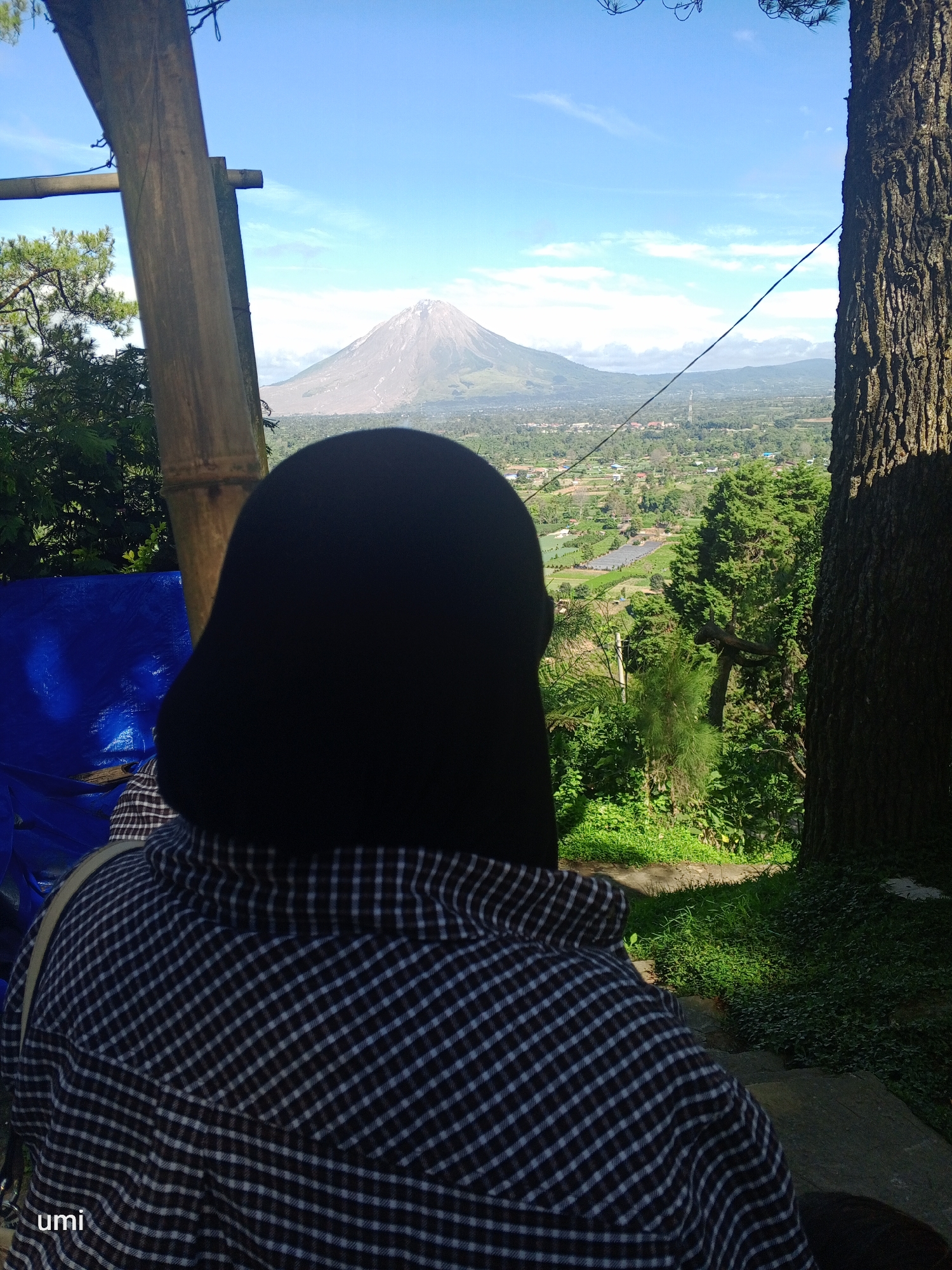 In the city of Gundaling 2, Indonesia, a person wearing a black hijab and a checkered shirt is seen from behind, looking out at a panoramic view of a valley and a distant volcano under a clear blue sky with scattered clouds. The person is positioned on a slightly elevated area, with a large tree trunk to their right and bamboo structures to their left. The foreground includes a blue tarp and dense green vegetation. The valley below shows a patchwork of fields and buildings, suggesting a rural or semi-urban landscape. The volcanic mountain, with its conical shape and visible slopes, is the dominant feature in the background. The lighting suggests it is daytime, with sunlight casting shadows on the ground and vegetation. A watermark "umi" is visible in the bottom left corner of the image.