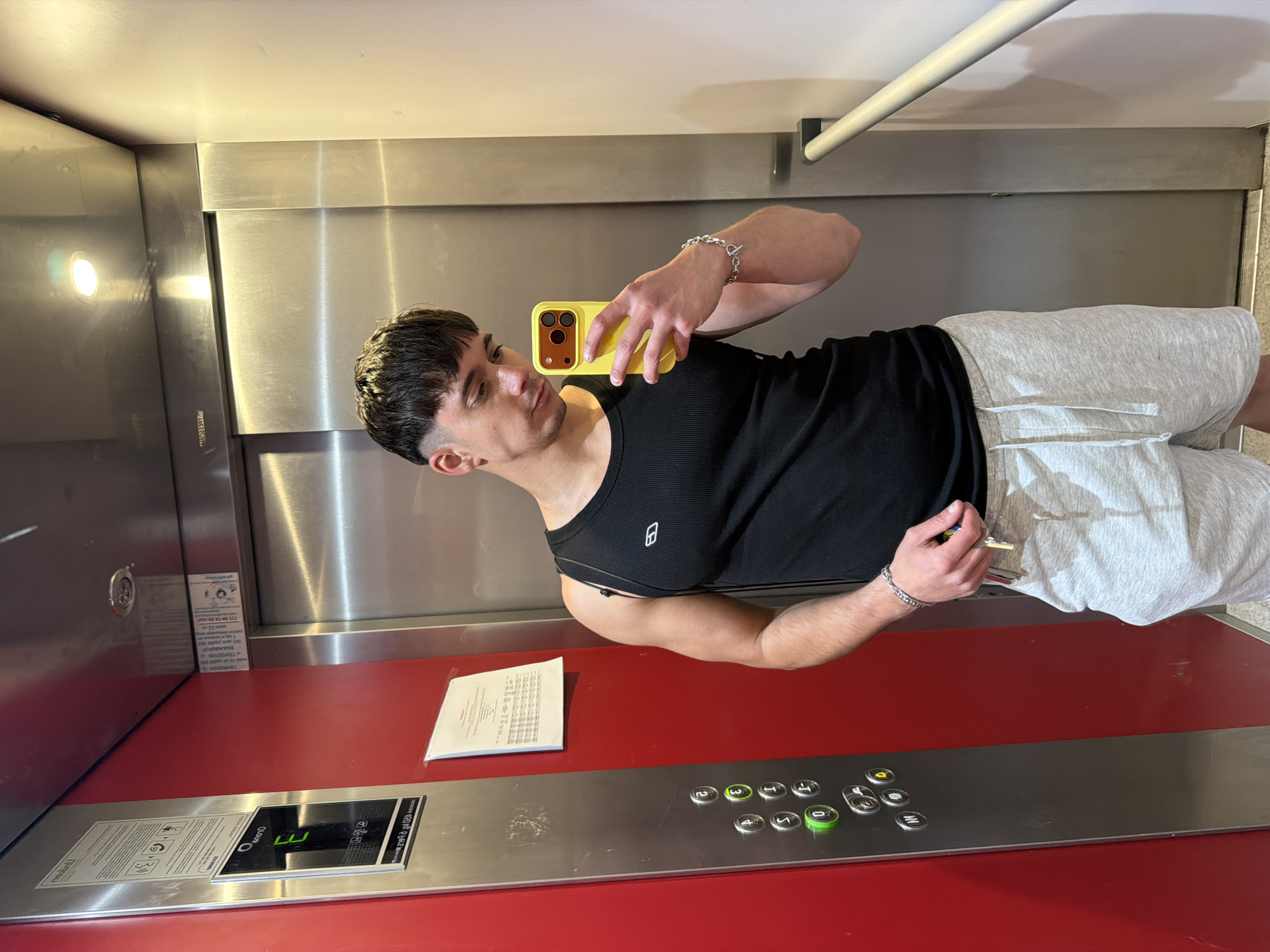 A young adult male stands in an elevator with red and stainless steel interior. He is taking a selfie with a yellow phone, wearing a black tank top and grey shorts. The elevator's control panel shows buttons numbered 0, 3, 1, 2, 4, and a display panel. A piece of paper is affixed to the red wall. The location cannot be determined to be in Sofia, Bulgaria from this image alone.