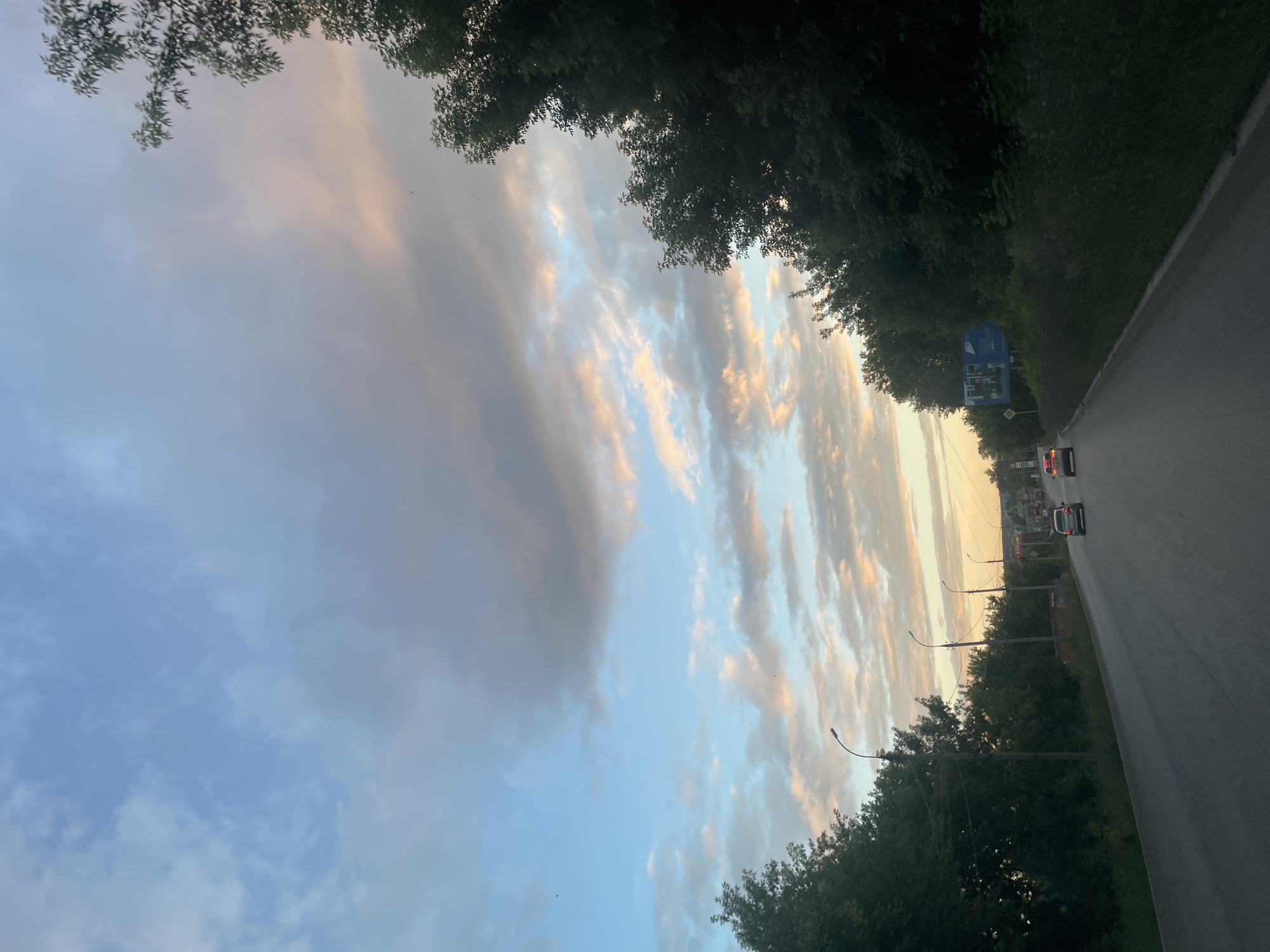 The image depicts a scenic view of a road in Votkinsk, Russia, during a picturesque sunset. The road, paved with asphalt, curves gently to the right, bordered by lush green trees and vegetation. Several cars are visible on the road, suggesting ongoing traffic.
The sky is a prominent feature, with a dramatic display of clouds illuminated by the setting sun. Streaks of orange, pink, and yellow are visible among the softer blues and grays of the clouds, creating a beautiful, atmospheric scene. The time of day is clearly sunset, as indicated by the warm hues in the sky.
Streetlights are spaced along the side of the road, some of which are illuminated, hinting at the transition from dusk to night. In the distance, a blue billboard with white text and images is visible, along with a smaller yellow diamond-shaped sign, likely a road warning sign. The overall mood of the image is serene and peaceful, capturing the beauty of nature as day turns into night.
