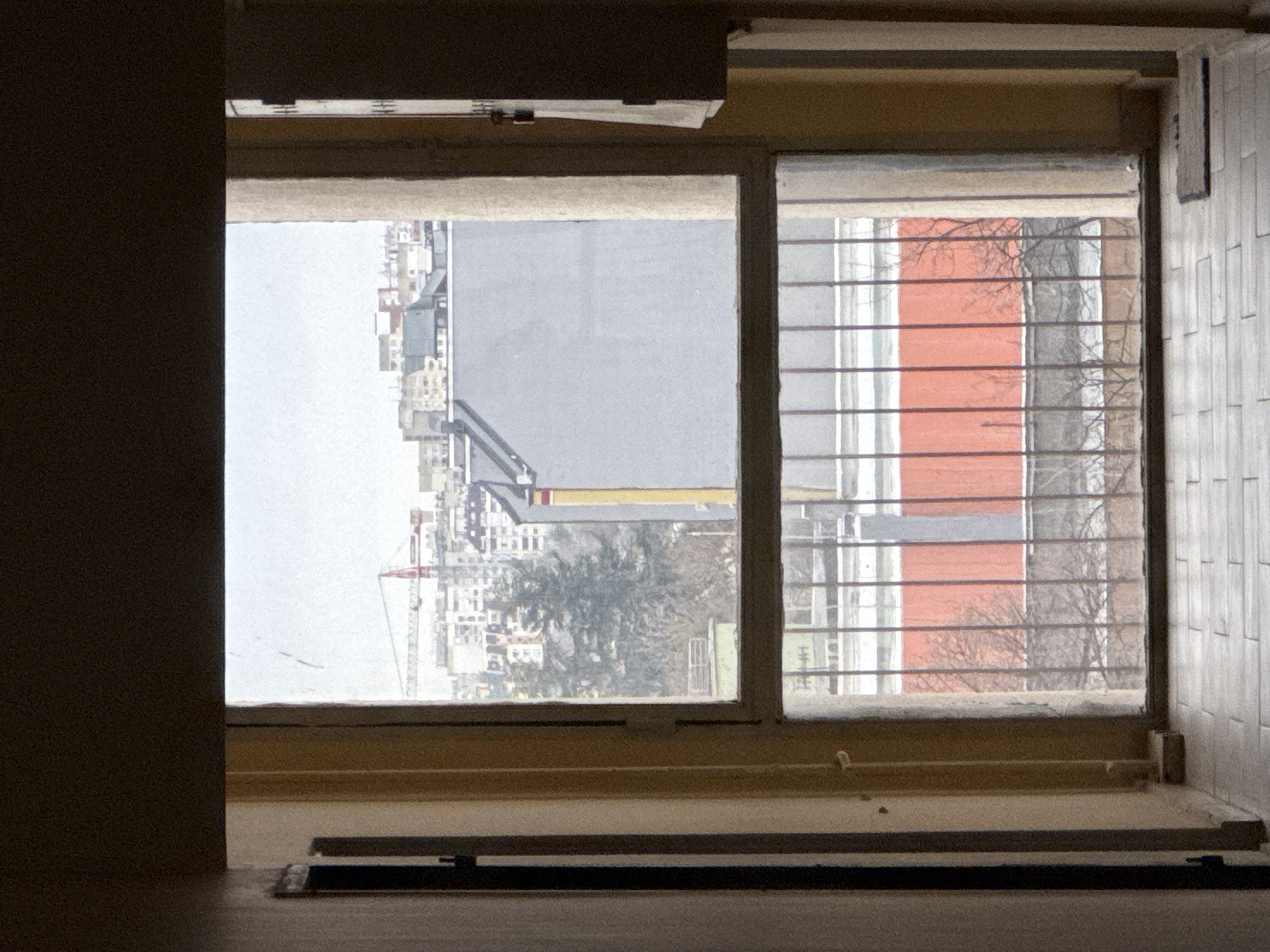 The image shows a view through a window. Outside, there's a cityscape. A building with a red facade is visible, partially obscured by a tree without leaves. There is a construction crane visible as well as dense urban buildings. The window has a security grate on the right pane. The window frame is beige. Below the window, there is a dark-colored object placed on the window sill. Given the style of the buildings and the overall urban environment, the scene could be located in Sofia, Bulgaria.