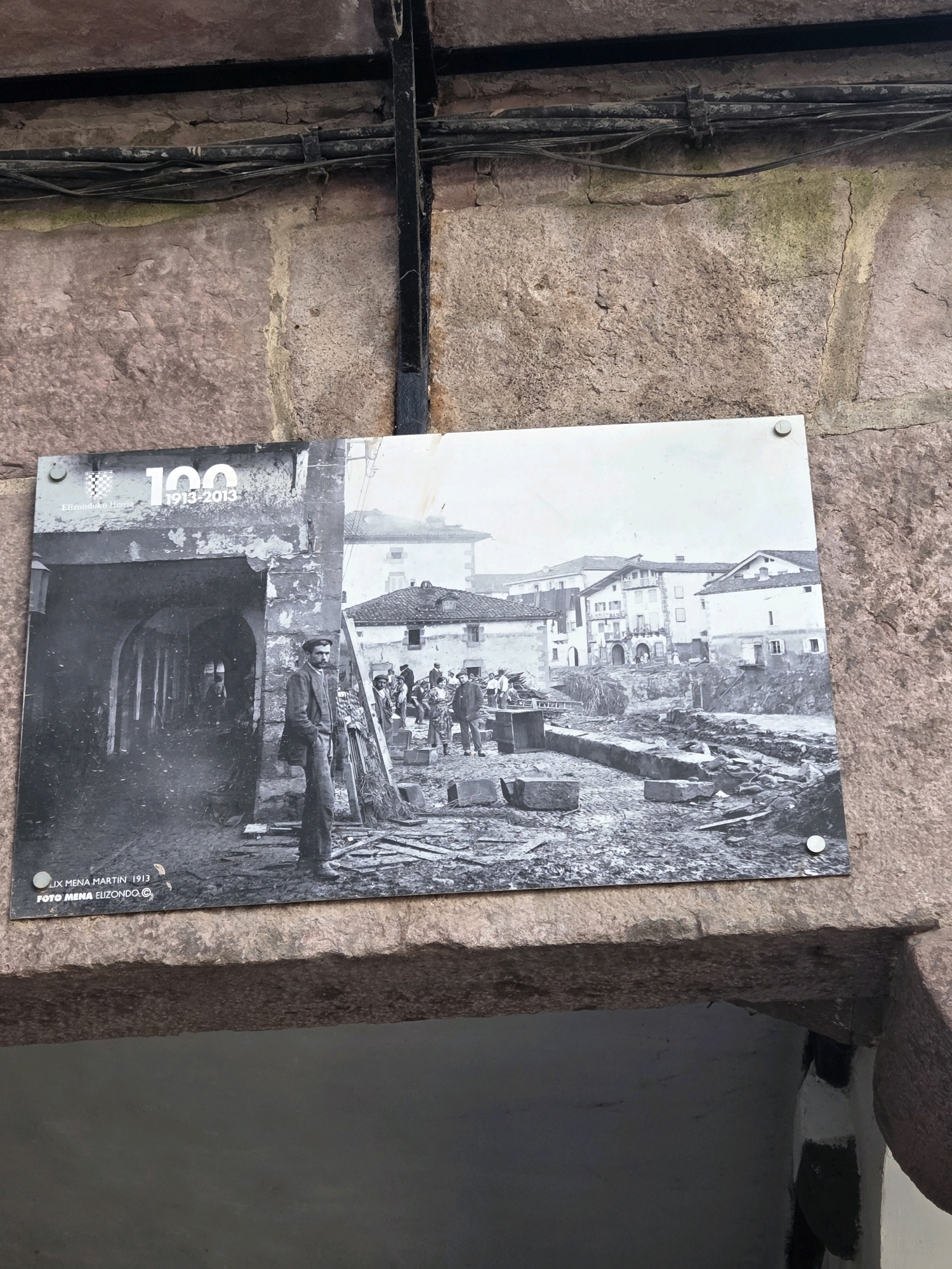 This black and white photograph is mounted on a stone wall, likely as a historical marker. The photograph depicts a scene from Elizondo, Baztan, Spain, with a prominent inscription "100 1913-2013" at the top left. The image shows a man standing in the foreground, dressed in a suit and hat, facing left. Behind him, an archway leads into a dark interior. To the right of the man, a street or plaza is visible, with rubble and debris scattered across the ground. Several other individuals, dressed in similar period clothing, are present in the background, some appearing to be engaged in activity. Buildings line the background, some with multiple stories. The bottom of the photograph includes text that reads "JUX MENA MARTIN 1913" and "FOTO MENA ELIZONDO.C.". The photograph is secured to the wall with visible metal fasteners. The surrounding stone architecture suggests an older building.