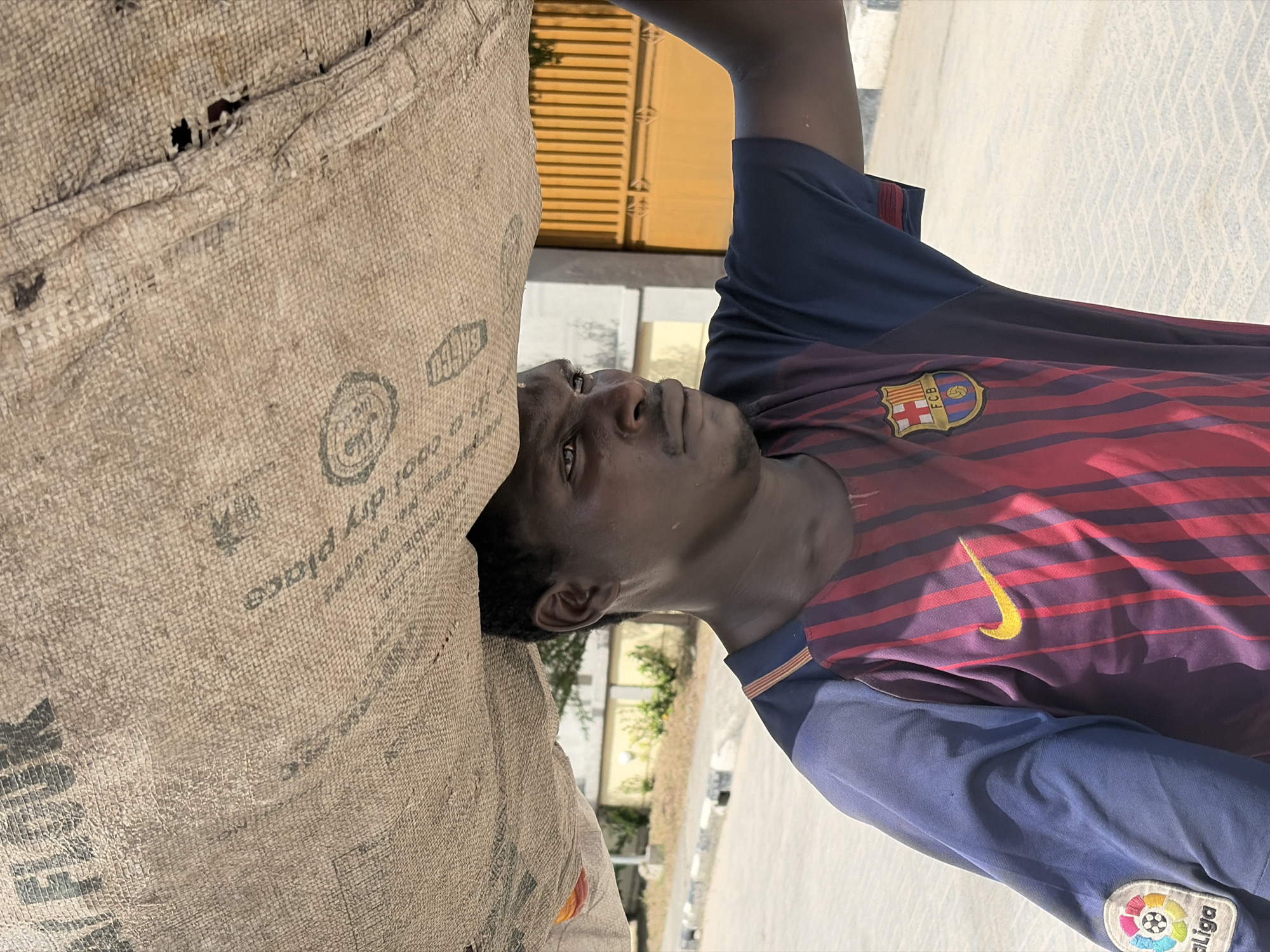 A young man in a FC Barcelona soccer jersey leans against a large woven bag. The location is Igboefon, Nigeria. The bag has a hole and text that includes "FLOUR" and "dry place". The background shows a street, sidewalk, and buildings.