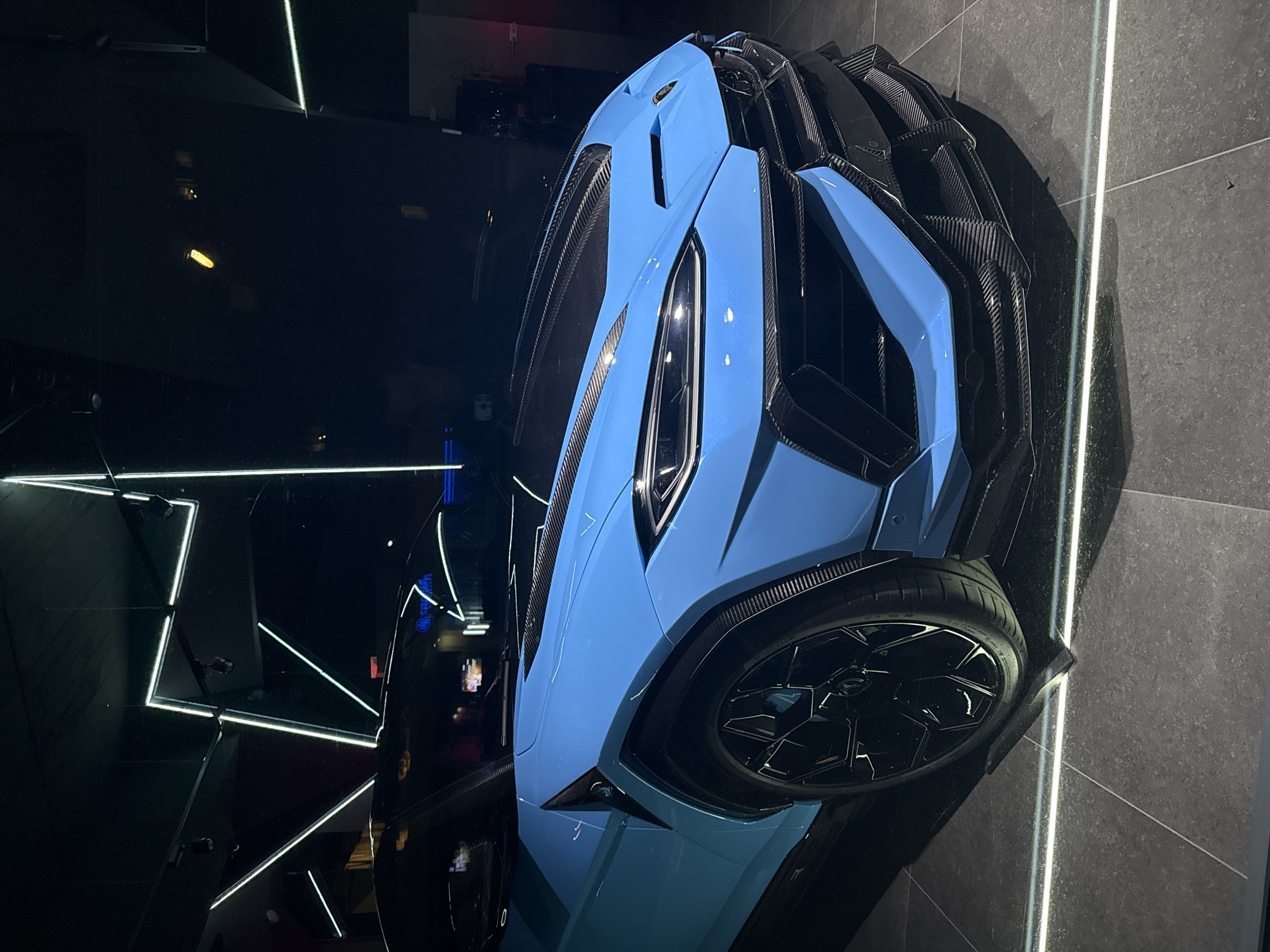 The image shows a bright blue Lamborghini Urus Performante parked indoors on a dark tiled floor. The car is positioned at an angle, with its front facing the viewer. The car features carbon fiber accents on the hood, front bumper, and wheel arches. The interior lighting appears to be a mix of white and neon green lines, suggesting a showroom or exhibition space. The background is dark and indistinct, with some reflections visible on the car's surface. There is no indication of the city of Munich, Germany, in the image.