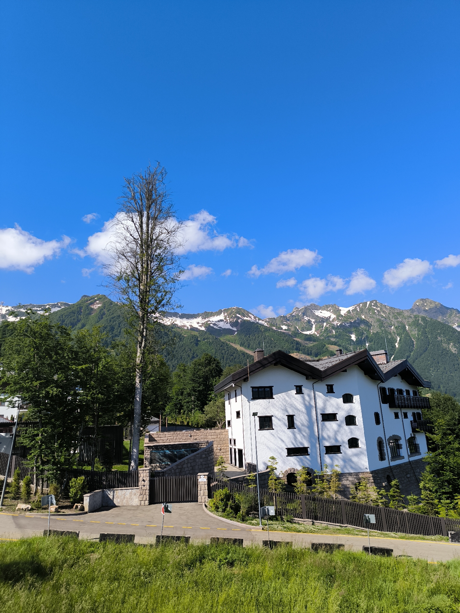This is a wide-angle, eye-level shot of a mountain resort scene in Estosadok, Russia, on a clear, sunny day. The sky is a vibrant blue with scattered white clouds. In the foreground, lush green grass and foliage line a paved road with yellow lane markings and concrete barriers. A black metal fence runs along the side of the road, separating it from the property.
Dominating the mid-ground is a large, white, multi-story building with a dark, sloped roof and numerous black-framed windows. The building appears to be a hotel or residential complex, built in a traditional alpine style with stone accents around the base. A black metal gate with stone pillars marks the entrance to the property.
Behind the building, a dense forest of green trees covers the lower slopes of the mountains. The upper parts of the mountains are rugged and rocky, with patches of snow still visible on the peaks. A tall, bare tree stands prominently in the center of the frame, reaching towards the sky. The overall impression is one of natural beauty combined with a well-developed tourist destination.