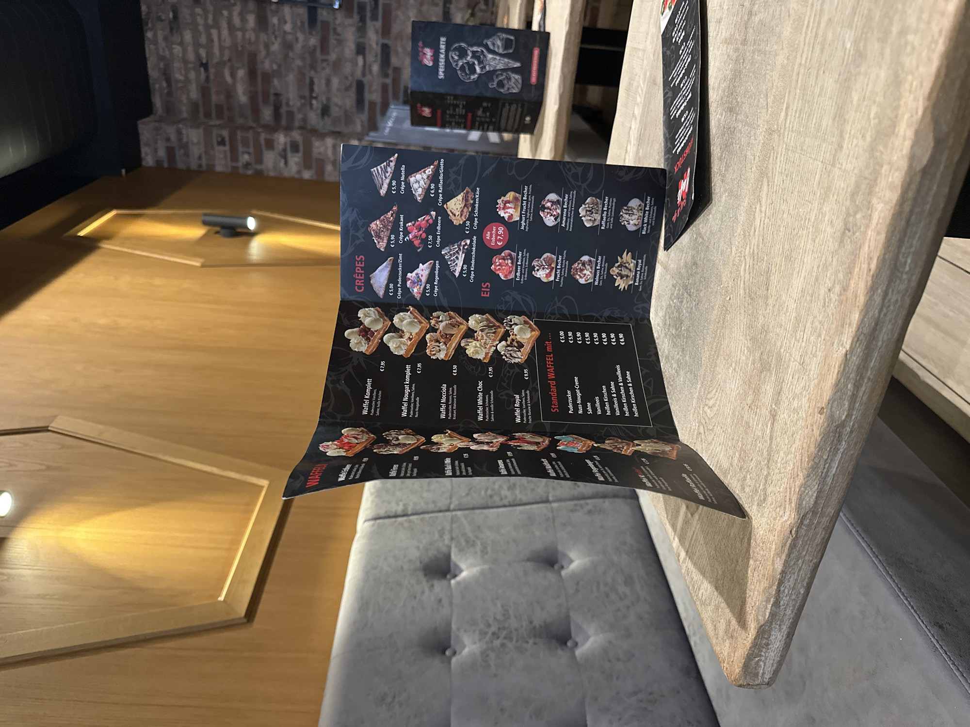 The image shows a menu on a wooden table inside a cafe in Krefeld, Germany. The menu is for waffles and crepes, and it is open to a page displaying various waffle and crepe options with prices. There are also ice cream options displayed. The cafe has a modern interior with a gray upholstered bench seat and wooden wall paneling. The lighting in the cafe is warm and inviting, with a spotlight illuminating the wall paneling. The overall impression is one of a cozy and pleasant dining experience.
