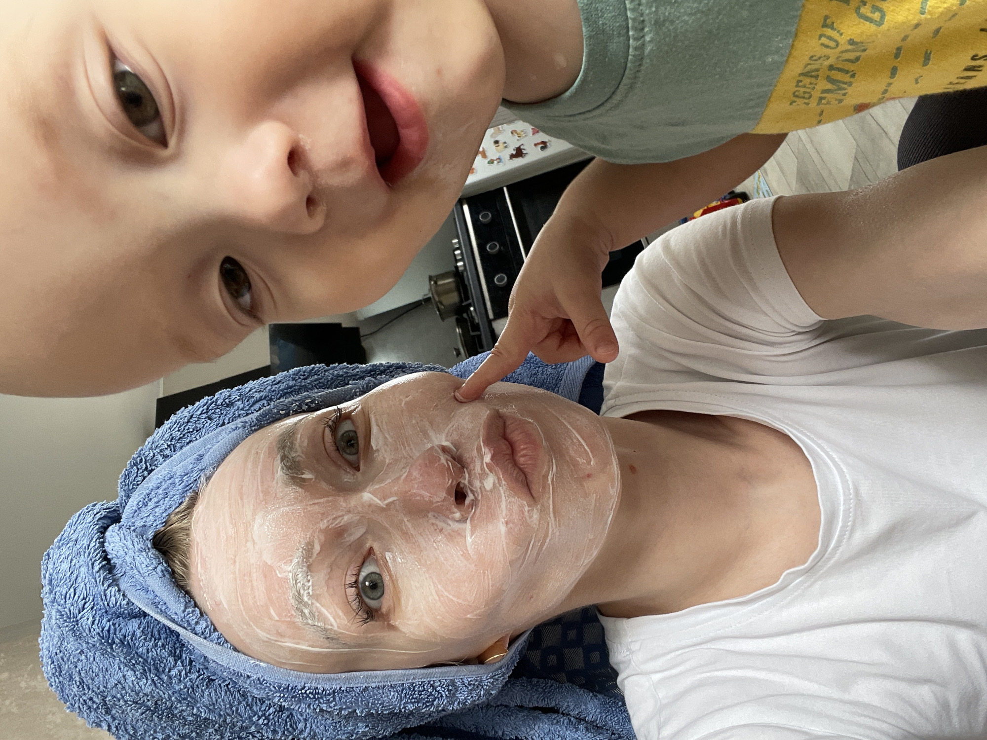 A selfie taken indoors in what appears to be a home setting. A young child with light brown hair is partially visible at the top of the frame, positioned to the left. The child is pointing their finger at a woman, presumably the child's mother. The woman's face is covered with a white facial mask and her hair is wrapped in a blue towel. She is wearing a white t-shirt.
In the background, a kitchen setting is vaguely visible, including what looks like a stainless steel stove. The location is identified as Perm, Russia, though specific visual details confirming this location are absent from the image.