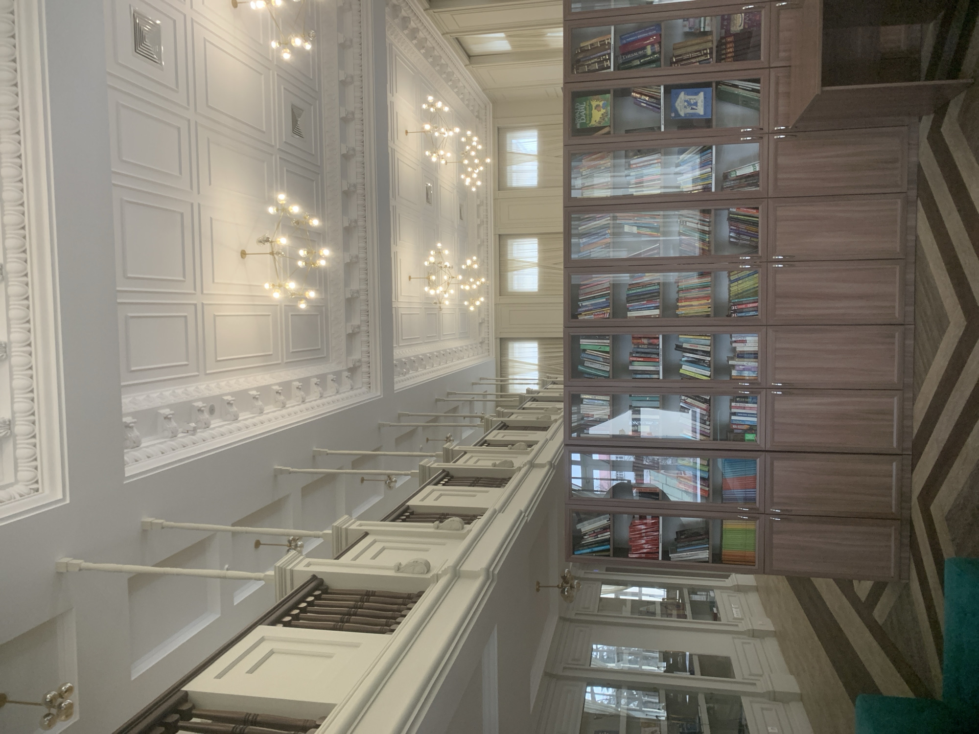 The image shows the interior of a room with high ceilings, likely a library or a similar space. The focal point is a large wall of bookshelves, filled with books and covered with glass doors. To the left of the bookshelves, the wall is white and decorated with detailed molding and evenly spaced light fixtures. Windows appear in the upper center of the frame. The image has been rotated or appears to be shot from an unusual angle. Based on visual clues and architectural styles, it is impossible to determine the specific location in Izhevsk, Russia, without additional context.