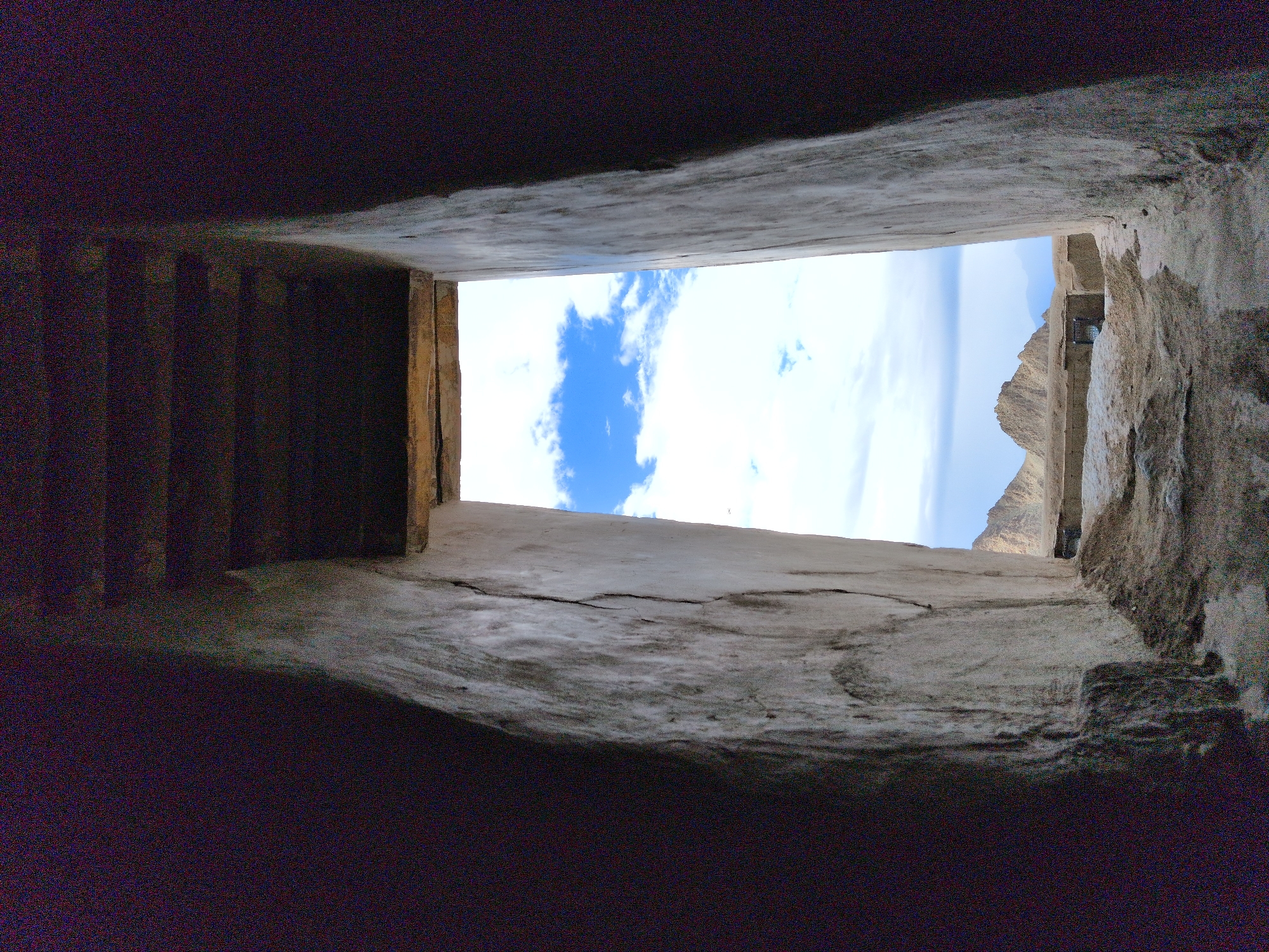 The image shows a view looking out through a rectangular opening in a stone structure. The opening frames a landscape with mountains in the distance under a blue sky with white clouds. The interior of the structure is dark and appears to be made of rough stone walls and a wooden ceiling. The ground in the foreground of the opening is covered in sand and loose rocks.