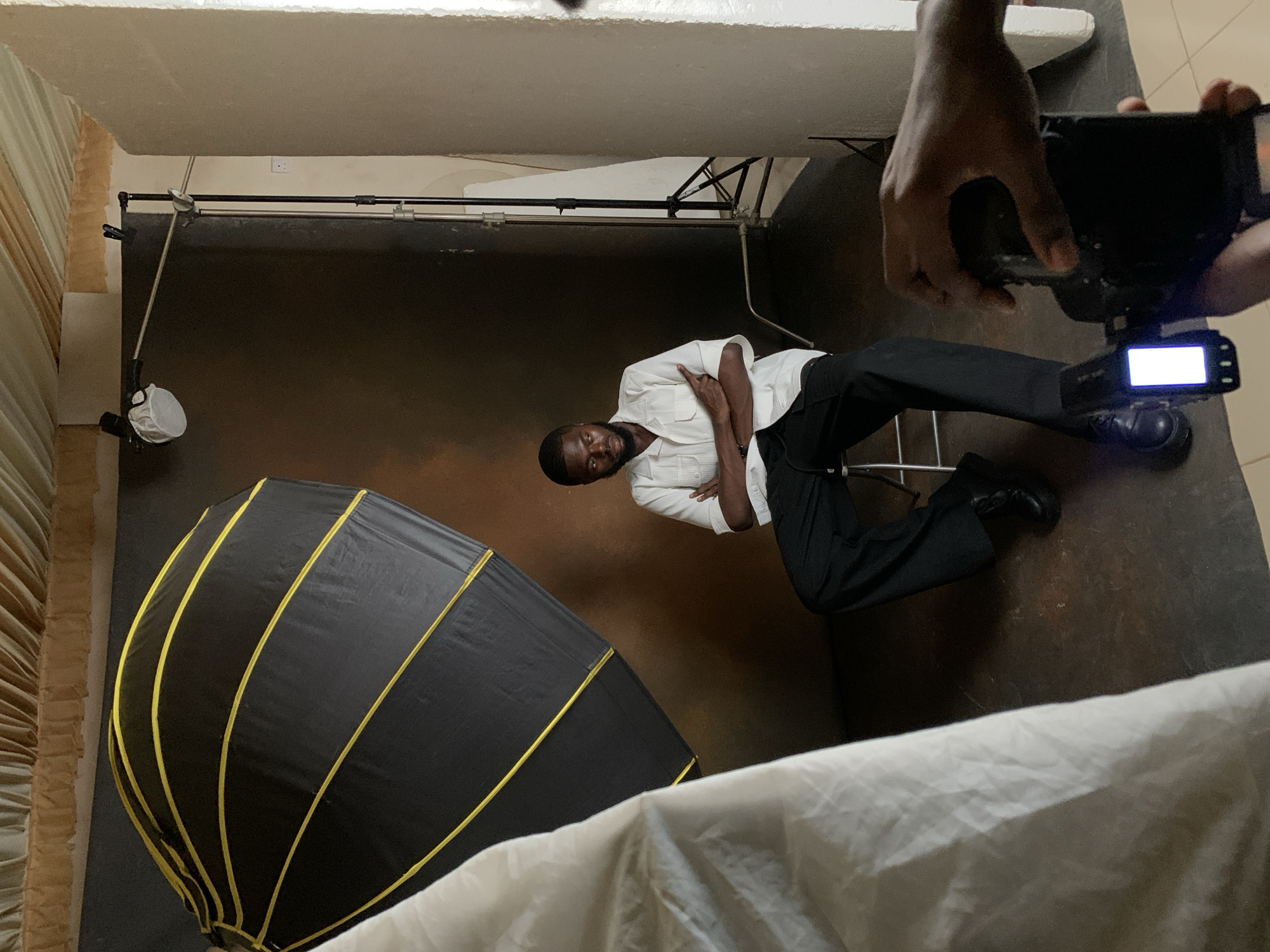 A young Black man with a beard sits with his arms crossed on a metal stool in front of a dark brown backdrop. He is wearing a white short-sleeved button-down shirt and black pants. To his left, a large black umbrella-like light modifier with yellow accents is positioned. In the foreground, a person's hands are holding a black camera. The setting appears to be an indoor photography studio. The location is identified as Kwang, Nigeria.