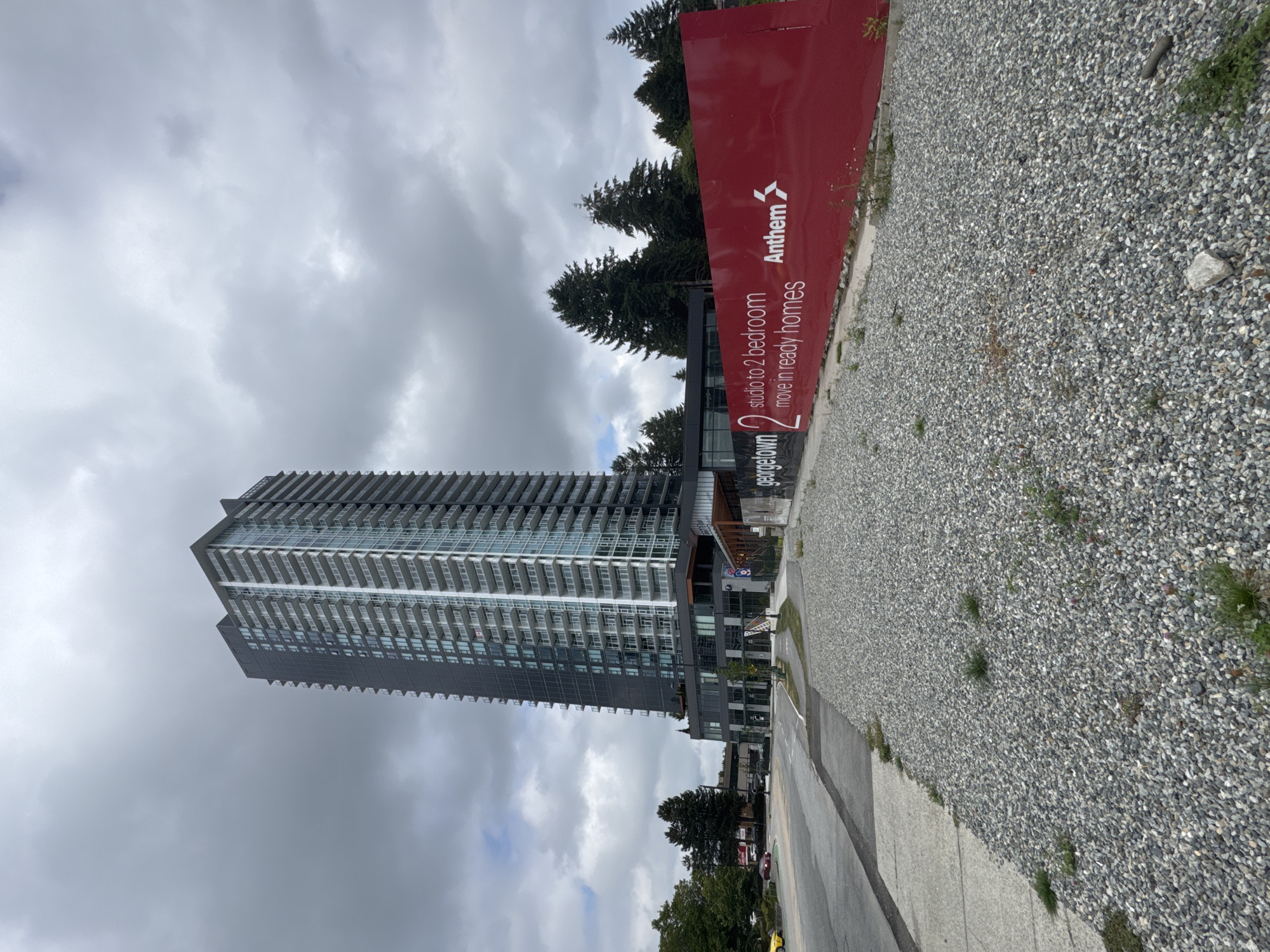 A wide, low-angle, exterior, daytime shot captures a modern, tall, residential building with a glass and dark-toned facade, looming under a cloudy, overcast sky. The building's numerous balconies are visible. In the foreground, a prominent, large, red billboard displays "georgetown 2 studio to 2 bedroom move in ready homes Anthem" in white text. The billboard is situated in front of a gravel-covered area. To the left, a paved road is visible, with cars driving on it. The scene appears to be part of a new development or construction site in Surrey, Canada, specifically within a likely urban or suburban area, given the presence of a major road and a large residential tower.