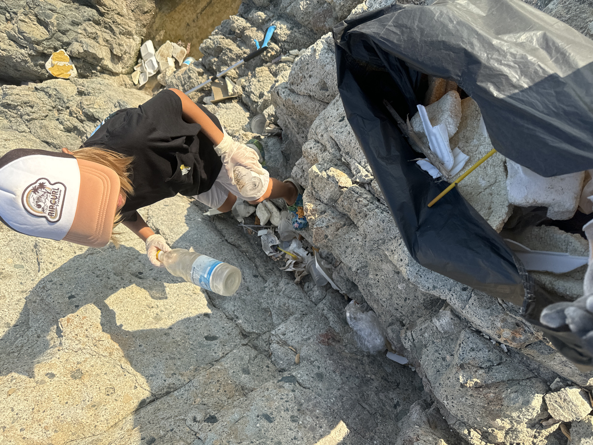 A person is picking up trash at a rocky beach in Mũi Né, Vietnam. The person, who appears to be a child, is wearing a black shirt, white shorts, and a hat with the Rip Curl logo. They are wearing gloves and picking up trash from the rocks and putting it into a large black trash bag. There is a clear plastic water bottle on the rocks next to them. The rocks are gray and jagged, and there is trash scattered among them. The trash bag is filled with styrofoam and other plastic waste. The sunlight is bright and casts a shadow of the person on the rocks.