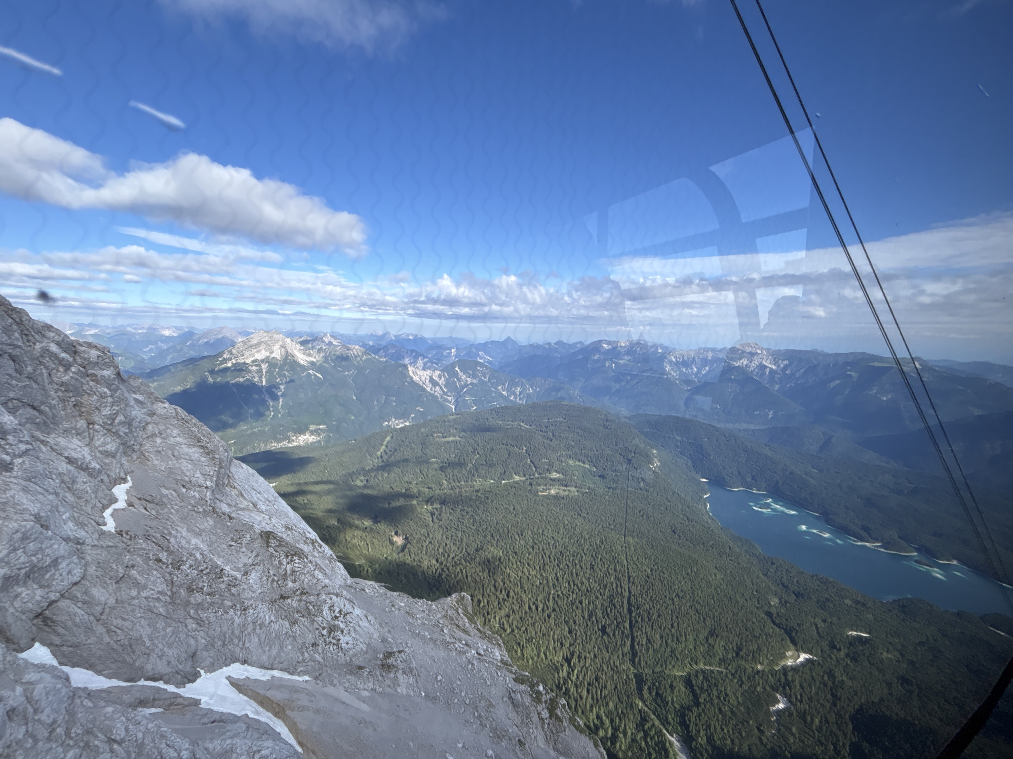 This image provides an aerial view of the Bavarian Alps, specifically the region surrounding Grainau, Germany, likely captured from a cable car. The foreground is dominated by a steep, rocky mountainside with patches of snow. In the mid-ground, a vast, dark green forest covers the rolling hills. To the right, a large, turquoise lake, possibly the Eibsee, is visible, nestled between tree-covered slopes. The background consists of a series of hazy, blue mountain ranges under a bright blue sky dotted with white clouds. Two dark cables, indicative of a cable car system, stretch across the upper right portion of the frame, suggesting the viewpoint. The overall scene depicts a scenic, mountainous landscape with a lake and dense forestation.