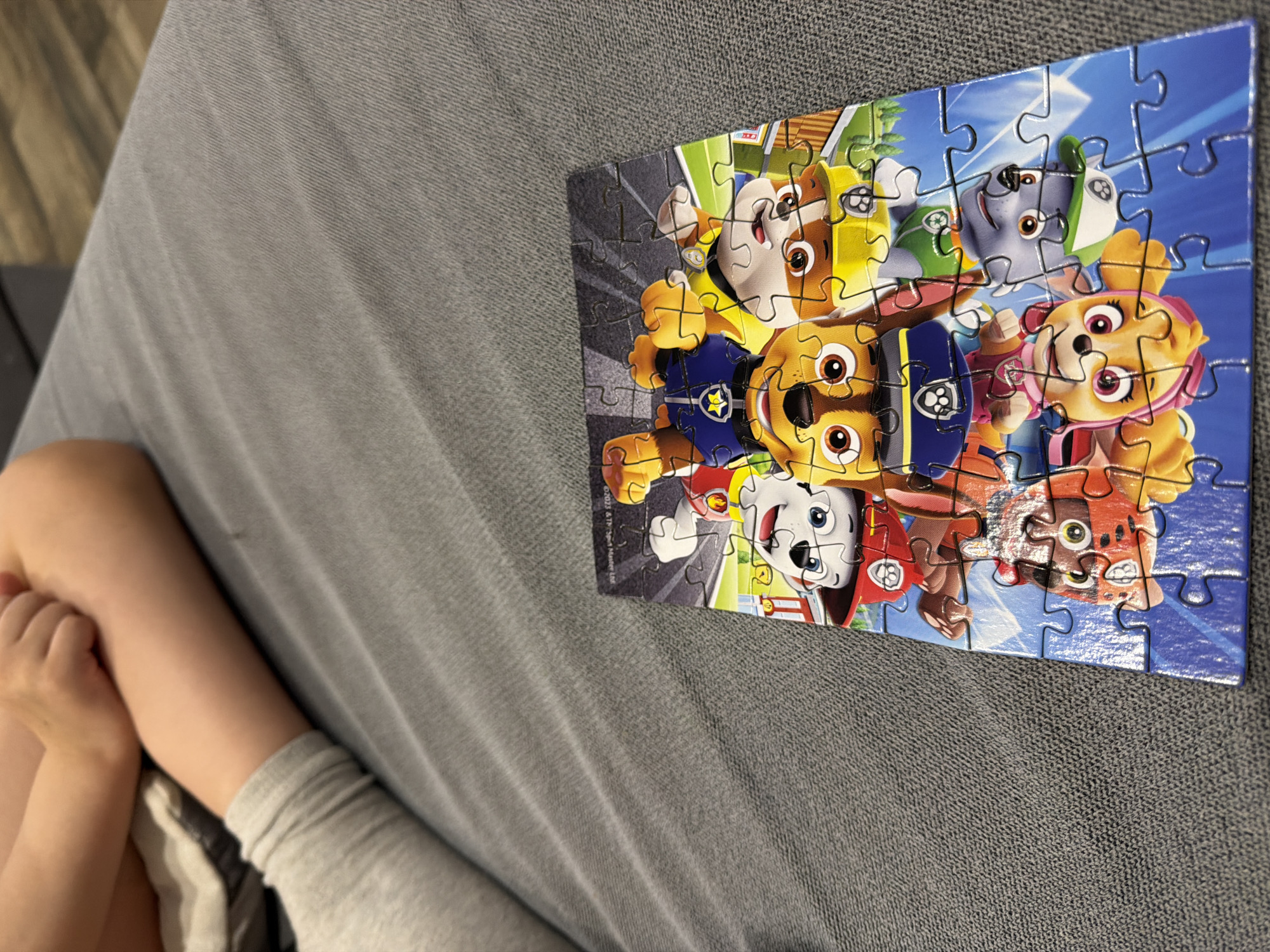 A partially completed Paw Patrol jigsaw puzzle, featuring multiple cartoon dog characters, sits on a textured gray surface, likely a sofa or blanket. A child's leg and part of their clothed body are visible on the left side of the frame. There is no discernible information in the image that relates to the city of Chaikovsky, Russia.