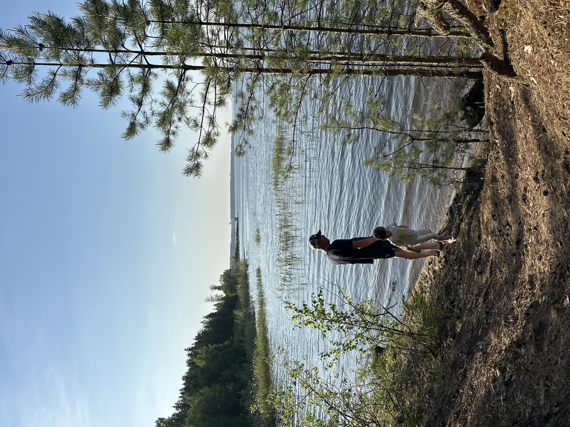 The image depicts a man and a child standing at the edge of a body of water. The man is wearing a dark shirt and a cap. The child is wearing a light-colored outfit. They appear to be standing on a sandy bank. In the background, there is a body of water, potentially a lake or a wide river, and a treeline on the opposite shore. Pine trees are visible along the shoreline near the individuals. The sky is clear and blue. The location is Chaikovsky, Russia.