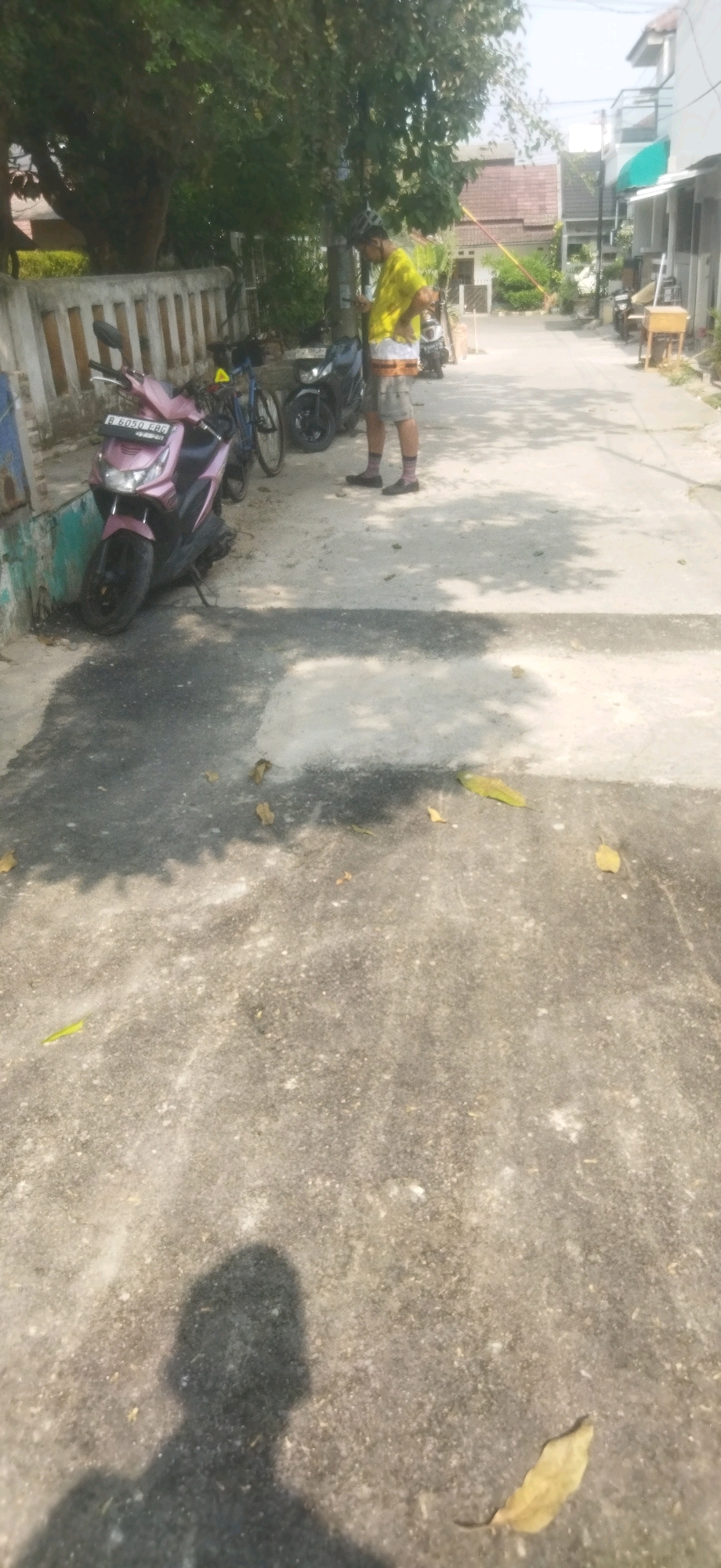 The image shows a narrow, paved street in Depok, Indonesia, likely in a residential area. The street is lined with houses and trees. Several motorcycles and a bicycle are parked along the side of the road. A person wearing a yellow shirt, grey shorts, and pink socks is standing in the street, looking at their phone. The person is wearing a helmet, suggesting they may have been cycling or riding a motorcycle. The ground is paved with concrete and shows signs of wear and tear, with some patches of darker discoloration. Shadows from trees and buildings are cast across the street, indicating it is a sunny day. There are a few fallen leaves scattered on the ground.
