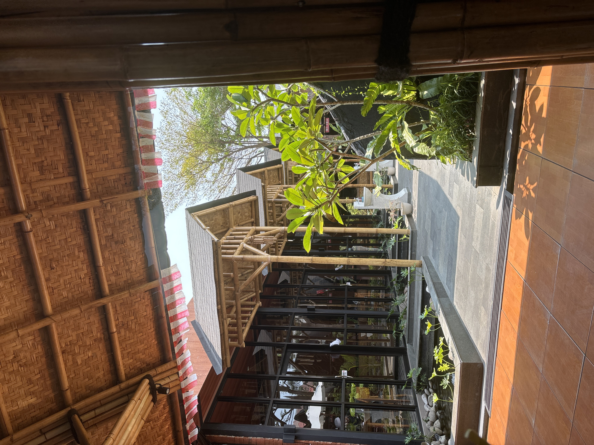 The image depicts an outdoor dining area with a bamboo-themed architecture in West Jakarta, Indonesia. The structure features a thatched bamboo roof and bamboo pillars, creating a rustic and natural ambiance. Large glass windows line the building, reflecting the surrounding greenery and sky. In the foreground, a small pond with aquatic plants and rocks adds to the serene atmosphere. Various potted plants and trees are strategically placed throughout the area, enhancing the lushness. The ground is paved with stone tiles in some sections and terracotta tiles in others, suggesting distinct zones within the outdoor space. The scene appears to be set during daytime, with sunlight casting shadows, indicating a clear sky. No people are visible in the image.