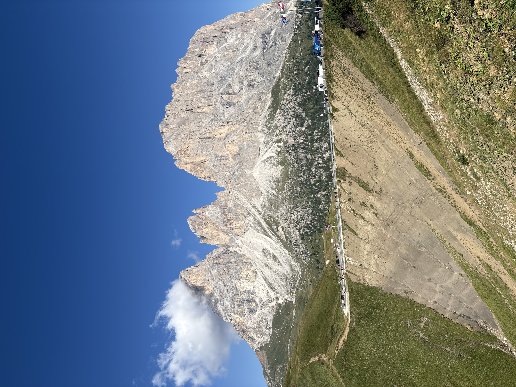 The image depicts a scenic mountain landscape under a clear blue sky. In the foreground, a steep, grassy slope descends into a rocky incline. A paved road runs along the middle ground, with several cars and people visible. Beyond the road, a dense forest of evergreen trees covers the lower slopes of the mountains. The prominent feature of the background is a massive, rugged mountain range with jagged peaks. Clouds partially obscure the upper portions of the tallest peaks. Several flags are visible near the road, suggesting a viewing point or a border crossing. The overall impression is a breathtaking view of the Italian Dolomites, likely near the city of Canazei, known for its dramatic mountain scenery and hiking trails.
