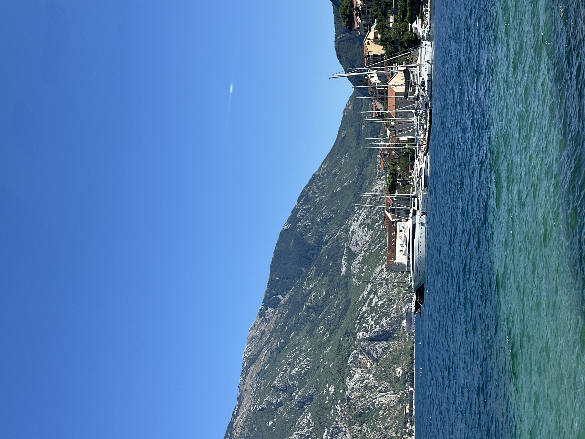 This is a photograph of the Bay of Kotor in Prčanj, Montenegro, on a clear, sunny day. The foreground is dominated by deep blue, rippling water, transitioning to a lighter, translucent turquoise closer to the bottom edge of the frame, revealing the seabed. In the midground, a marina is visible, filled with numerous sailboats with their masts pointed skyward. A large, white motor yacht is prominently docked. Behind the marina, a cluster of buildings with red-tiled roofs and light-colored facades are nestled on a hillside. The background features a large, steep, green-covered mountain with rocky outcrops, rising dramatically against a clear blue sky. A faint, wispy cloud is visible high in the sky. The overall scene depicts a serene coastal landscape with a mix of natural beauty and a developed harbor.