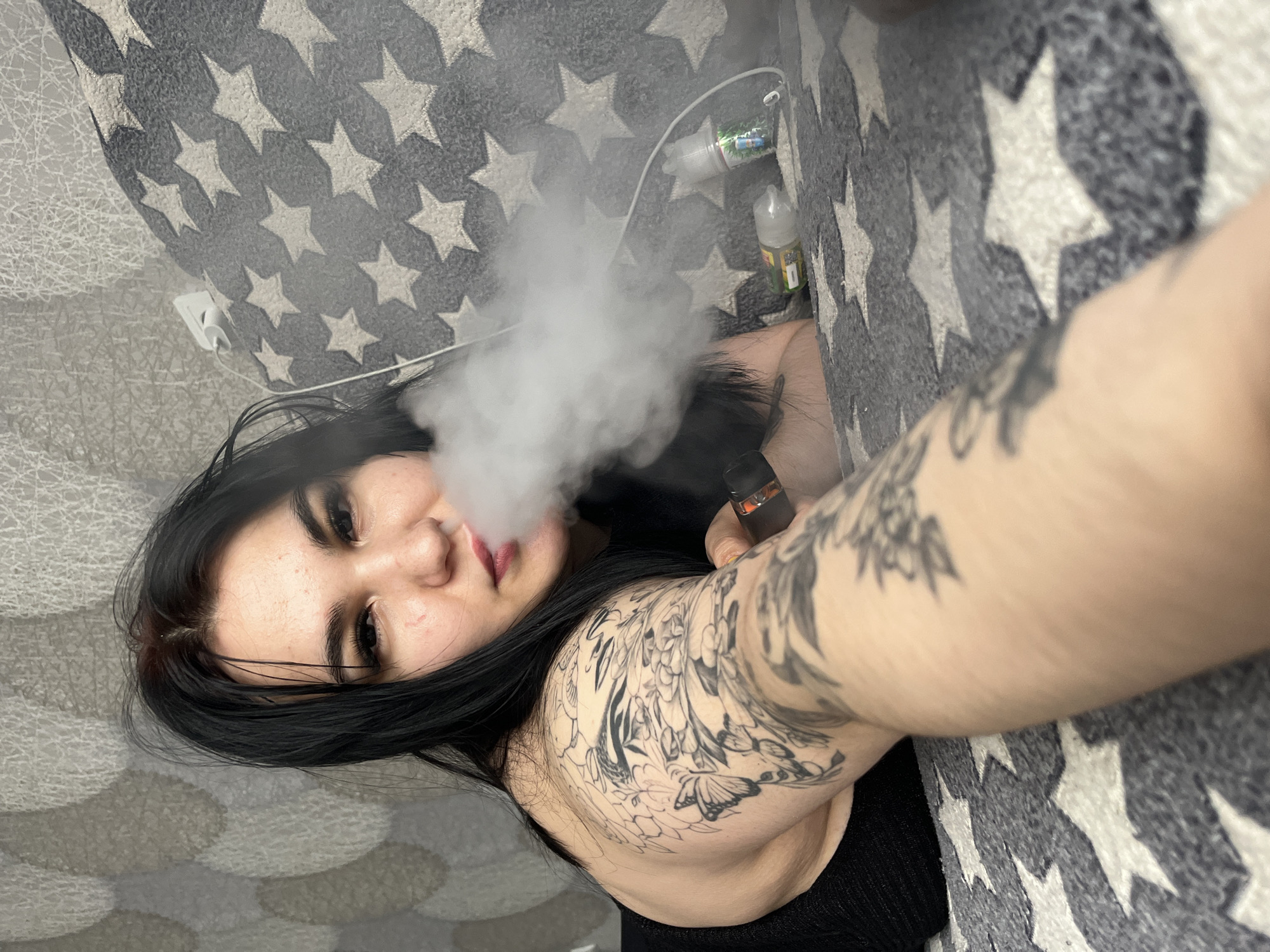A young woman with dark hair is vaping while lying on a bed with a gray star-patterned blanket. She has tattoos on her arm and shoulder. Vaping liquid bottles and a charger are located on the blanket near her head. The location is identified as the city of Chaikovsky, Russia.