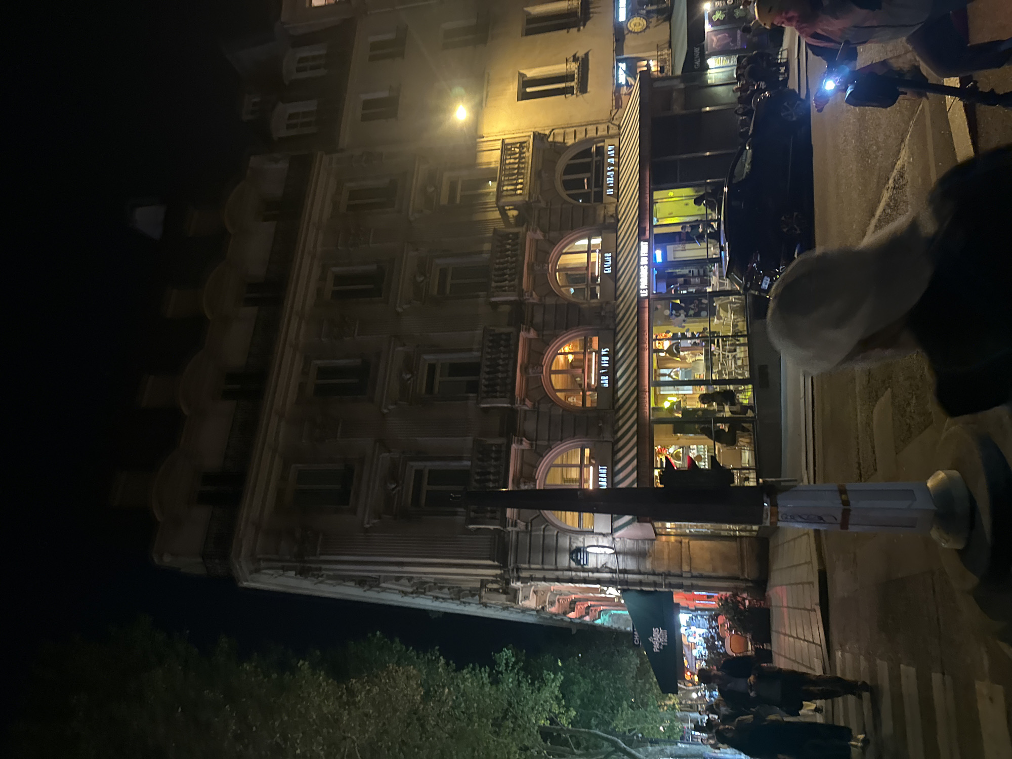 A street scene in Paris at night. A tall, ornate building with arched windows houses "Le Somas des Fruits" restaurant. Pedestrians are seen walking on the sidewalk and crossing the street. There is a parked car, and a person on a scooter with a headlight. There are trees in the foreground. The environment is dark but illuminated by street lights and building lights.