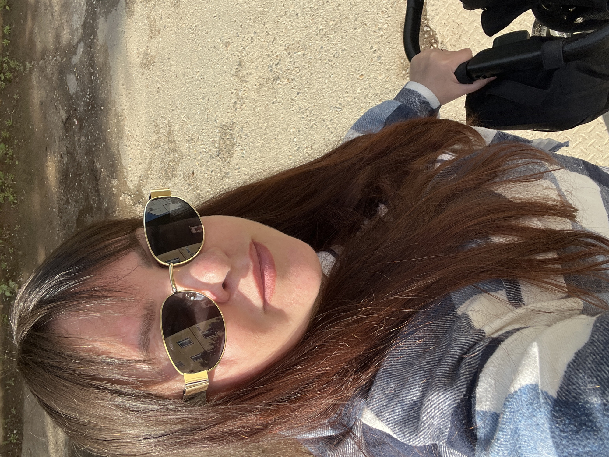 A woman wearing sunglasses and a blue and white plaid shirt is shown from an aerial perspective. She is holding the handle of a stroller. The background appears to be a paved surface. The reflection in the sunglasses indicates an urban environment, possibly including buildings.