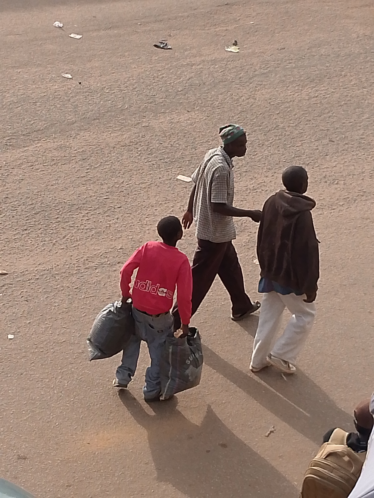 In Bukuru, Nigeria, three individuals are walking on a dusty, asphalt-like surface. In the foreground, a child wearing a pink "Adidas" sweatshirt and blue jeans carries two large, dark garbage bags. Behind the child, a man in a plaid shirt and dark trousers walks, with his right arm extended as if gesturing or reaching out. To the right of the man in plaid, another individual in a dark hooded jacket and white trousers walks. The ground is littered with small pieces of debris. A portion of a tan bag or backpack is visible in the lower right corner of the frame. The lighting suggests it is daytime, casting shadows on the ground.