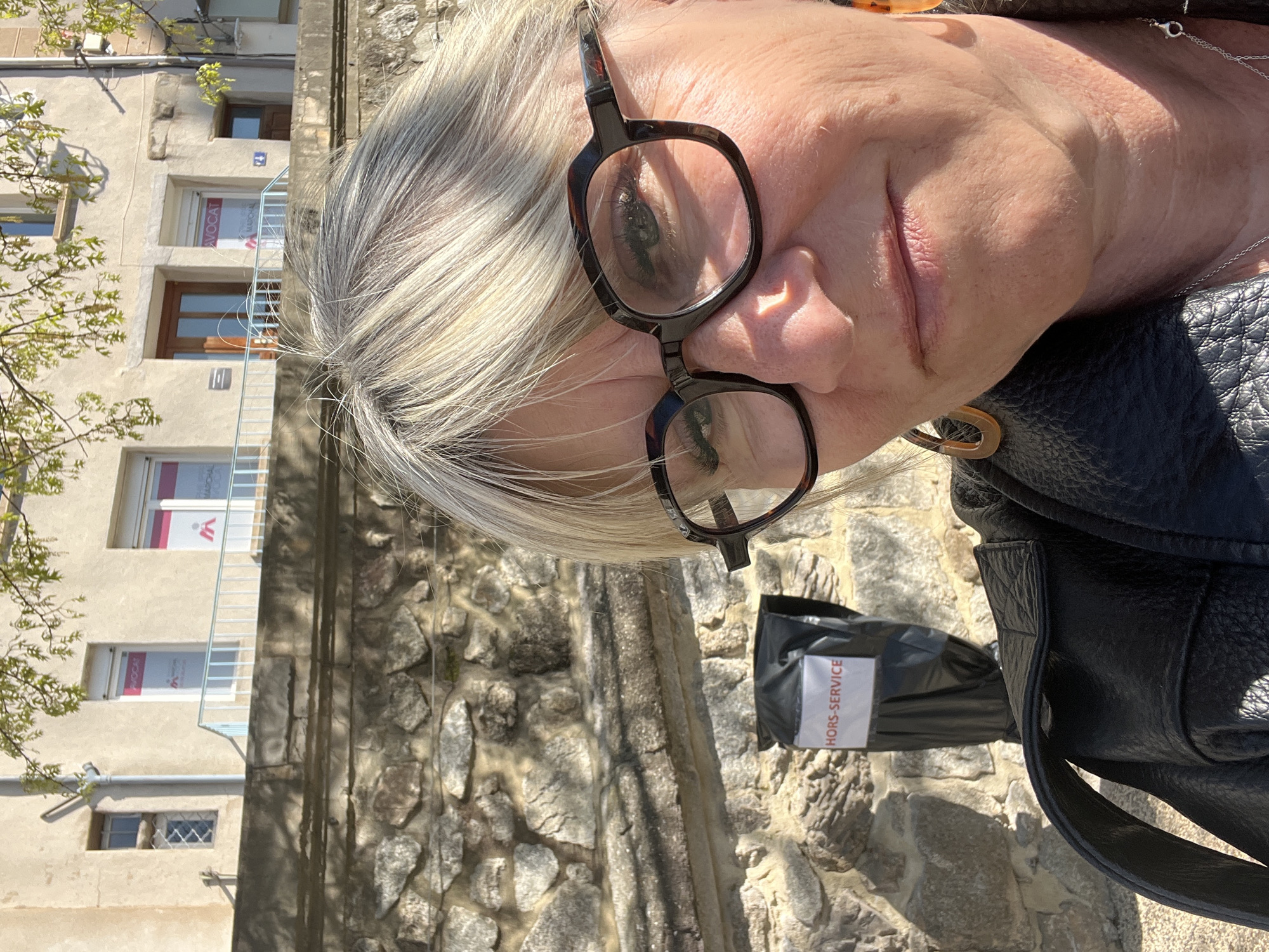 A close-up, off-center, eye-level shot shows a fair-skinned woman with short, gray-blonde hair and round tortoise-shell glasses looking directly at the camera. She is wearing a black leather jacket and a silver necklace.
Behind her is a stone wall, and further back is a beige building with several windows. The windows feature red and white signs labeled "AVOCAT" and what appears to be "MAZURKIE." A black bag marked "HORS-SERVICE" is attached to the wall to the woman's right.
The photo appears to be taken outdoors, likely in Saint-Galmier, France, given the request.
