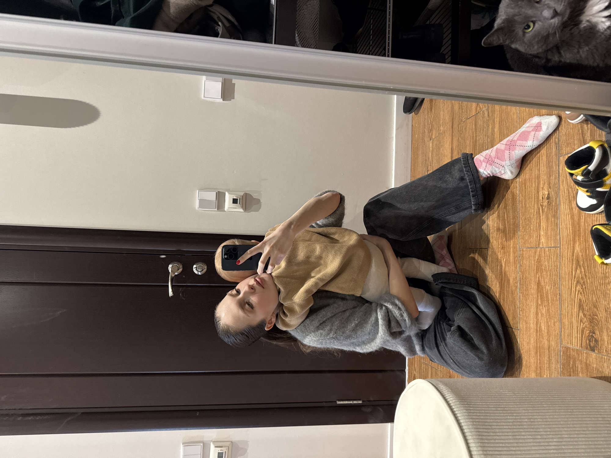 A woman is seated on the floor in what appears to be an apartment or home interior, based on the presence of a door, wall-mounted electrical outlets, and wooden flooring. She is holding a young child in her arms and taking a selfie with her smartphone. She is wearing dark jeans, a grey sweater, and patterned socks. The child is wearing a light brown top. The setting is indoors. The presence of the cat in the upper right suggests it is a domestic setting. Moscow, Russia, cannot be determined from this image.