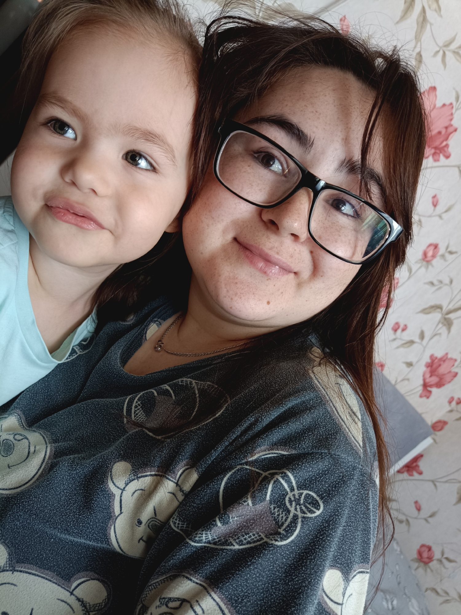 The image is a close-up, over-the-shoulder selfie of two individuals, a young child and an adult, against a floral wallpaper background. The child, positioned on the left and slightly in front, has light brown hair and is looking to the left with large, light-colored eyes. They are wearing a light blue garment. The adult on the right is wearing glasses with black frames and has brown hair. They are also wearing a dark blue garment with a pattern of cartoon bear faces. The adult is looking towards the camera and has freckles across their nose and cheeks. A thin silver necklace with a small pendant is visible on the adult's neck. The lighting suggests an indoor setting, likely a home environment. No specific location in a city is discernible from the image.