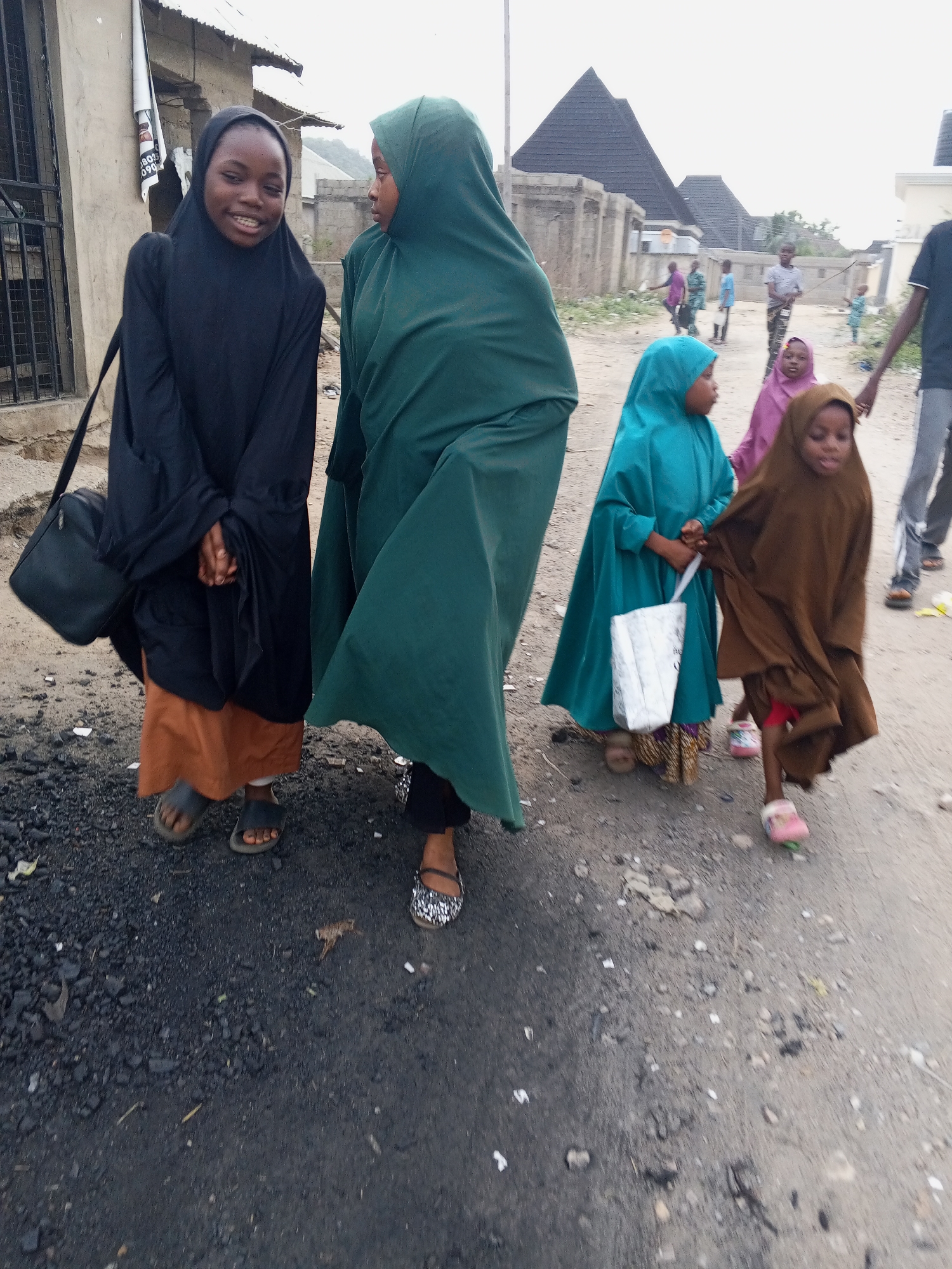 Otokiti, Nigeria, features a group of children walking on a partially paved road. Two taller children are in the front, one wearing a black hijab and a brown skirt while carrying a black bag; the other is wearing a green hijab and a long dress. Two smaller children are behind them, one wearing a turquoise hijab and dress, carrying a white bag, and the other wearing a brown hijab and dress. Other people are in the background. Some houses with pyramid-shaped roofs and other buildings are visible in the background. The ground is a mix of dirt, asphalt, and rocks.