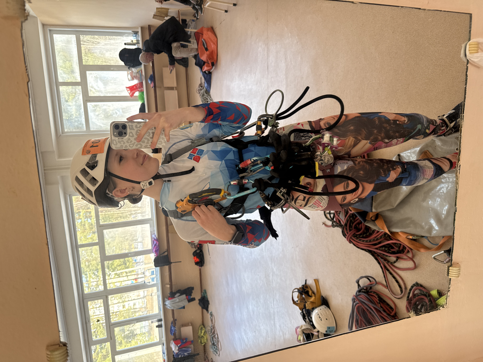A woman is taking a selfie in a mirror indoors. She is wearing a white climbing helmet with an orange sticker, a blue and white jacket, and patterned leggings depicting faces. She is also wearing a climbing harness with various carabiners and ropes attached. The mirror reflects a cluttered room with a large window showing trees and buildings, along with various items of clothing, ropes, and climbing gear scattered on the floor and benches. In the background, out of focus, are several people. The photo was taken in Usolye, Russia.