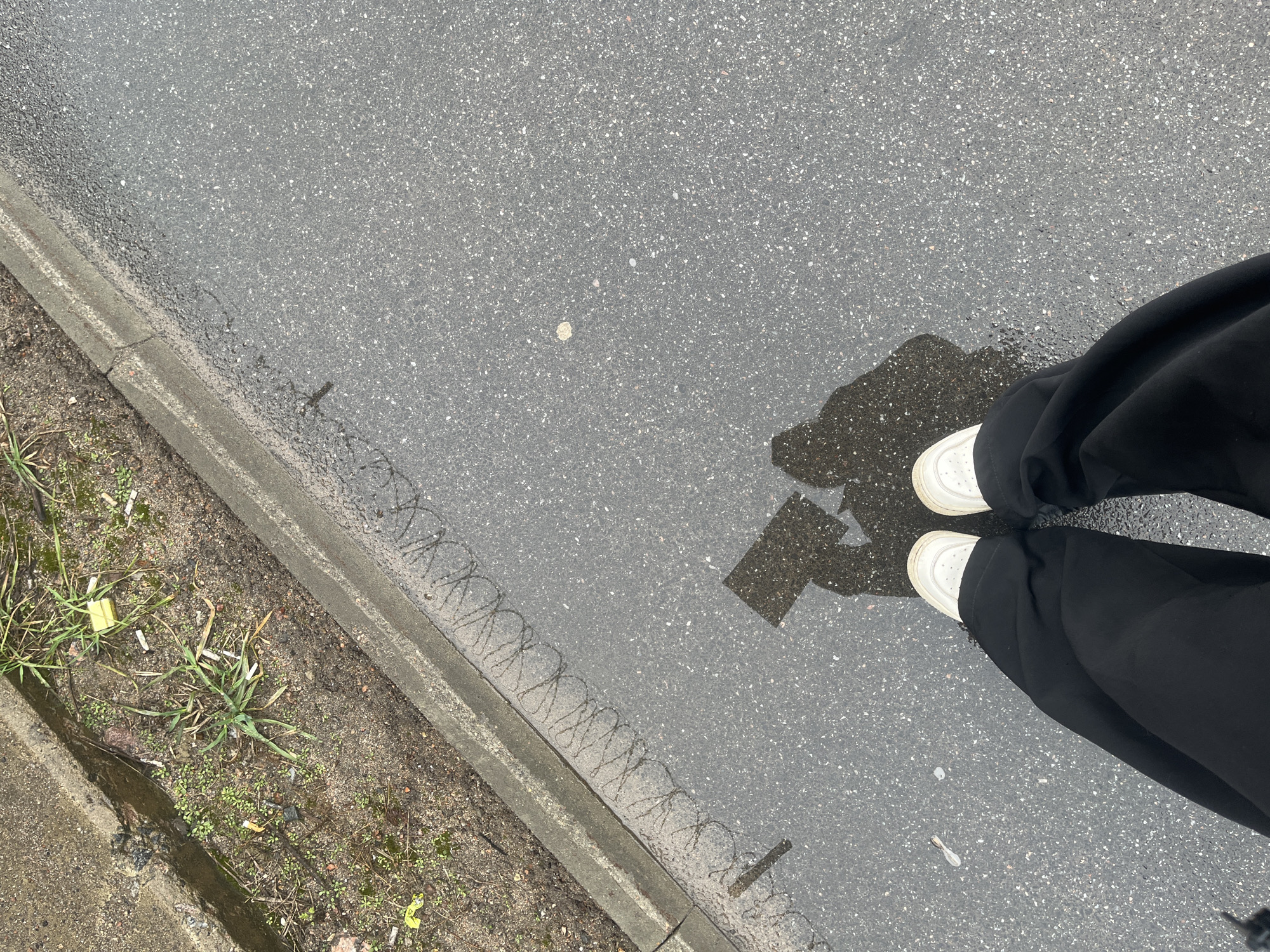 A top-down shot depicts a person standing on a wet, gray asphalt surface, likely a street or sidewalk. The person wears black, wide-legged pants and white sneakers. Their shadow is visible in a shallow puddle between their feet. To the left of the asphalt, a concrete curb separates the pavement from a patch of dirt with sparse green vegetation and litter, including discarded cigarette butts. A thin, linear accumulation of detritus is visible at the curb's edge, suggesting the recent presence of water flow. The image's location is unidentifiable as Saint Petersburg, Russia, without additional visual or contextual information.