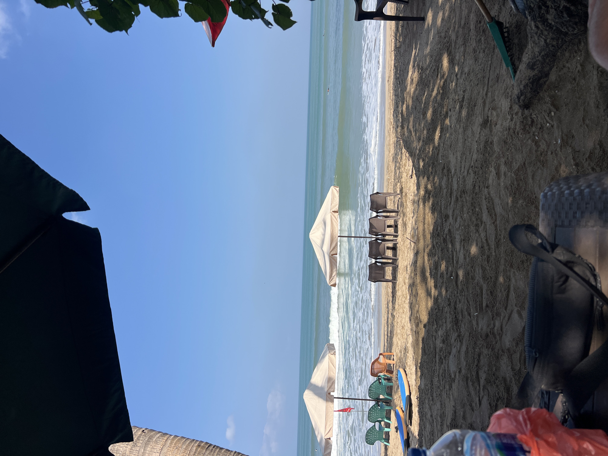A beach scene in Kuta, Indonesia, on a sunny day with a clear blue sky. In the foreground, a dark woven chair with a black bag and a plastic water bottle on a table. Further back, two surfboards are laid out on the sand. A row of plastic chairs, some green and some orange, sit near two beige beach umbrellas. Beyond the umbrellas, the ocean stretches out with gentle waves rolling onto the shore. In the distance, a person is visible in the water, possibly surfing. The sand is a light brown, with shadows cast from palm trees.