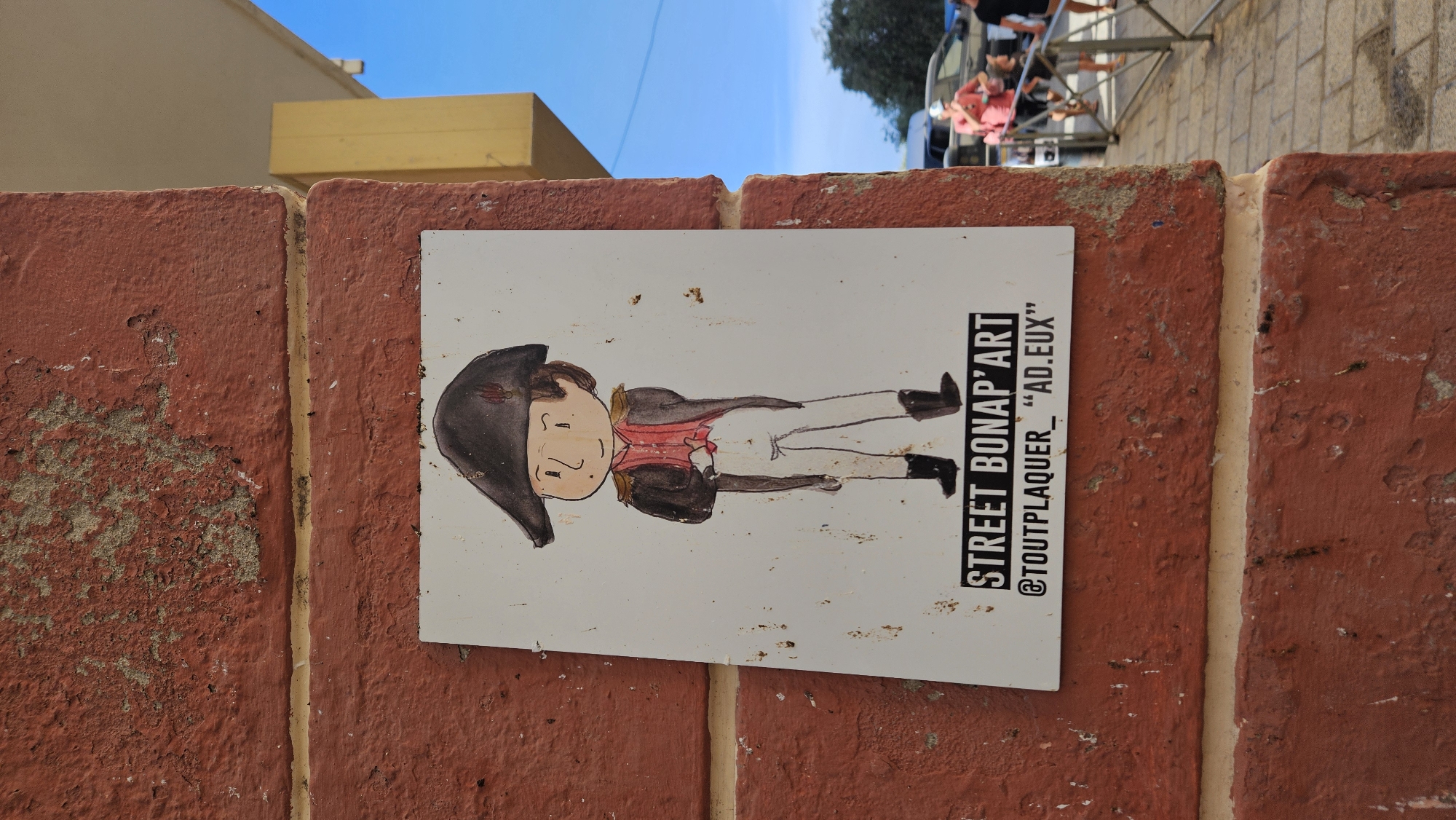 A photograph captures a piece of street art featuring a cartoonish depiction of Napoleon Bonaparte affixed to a red, weathered brick wall. The artwork, on a white rectangular surface, shows Napoleon in his characteristic bicorne hat, red waistcoat, white breeches, and black boots. Below the illustration, the text "STREET BONAP'ART" is emblazoned, followed by "@TOUTPLAQUER \"AD.EUX\"". The wall itself shows signs of age, with peeling paint and uneven texture. To the right of the artwork, a blurred background reveals a paved walkway, people in casual attire, vehicles, and a clear blue sky. This particular piece of street art is located on Rue Notre Dame in L'Île-Rousse, Corsica, France, known for its charming historical buildings and vibrant street art scene.
