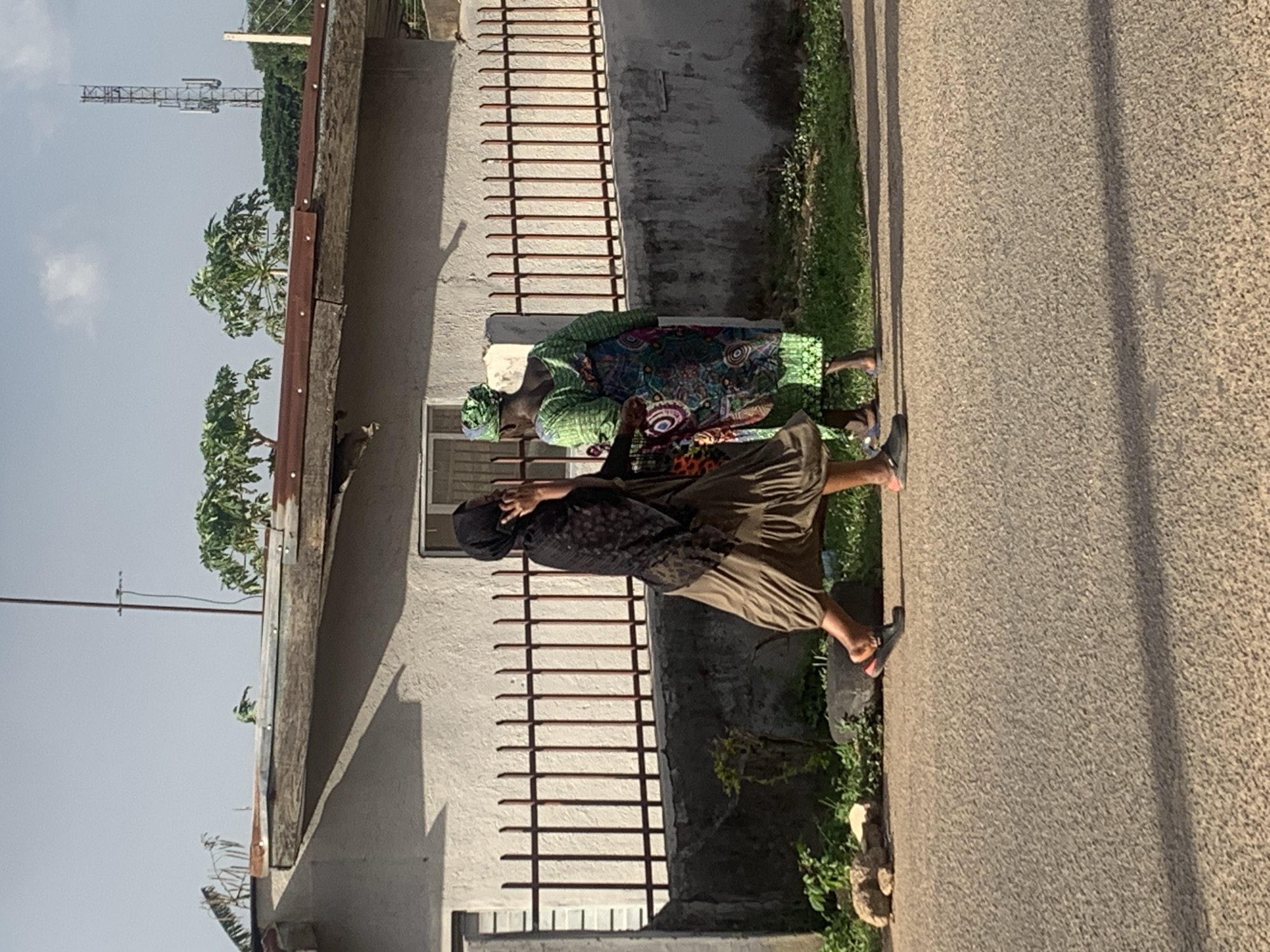 Two women stand on a paved road next to a building in Jos, Nigeria. The woman closest to the camera is wearing a black head covering, a black top with a pattern, and a brown skirt. The other woman wears a green and patterned dress with a matching head covering. The building next to them is white with a brown fence-like structure attached. Vegetation grows between the road and the building. A communication tower is visible in the background.
