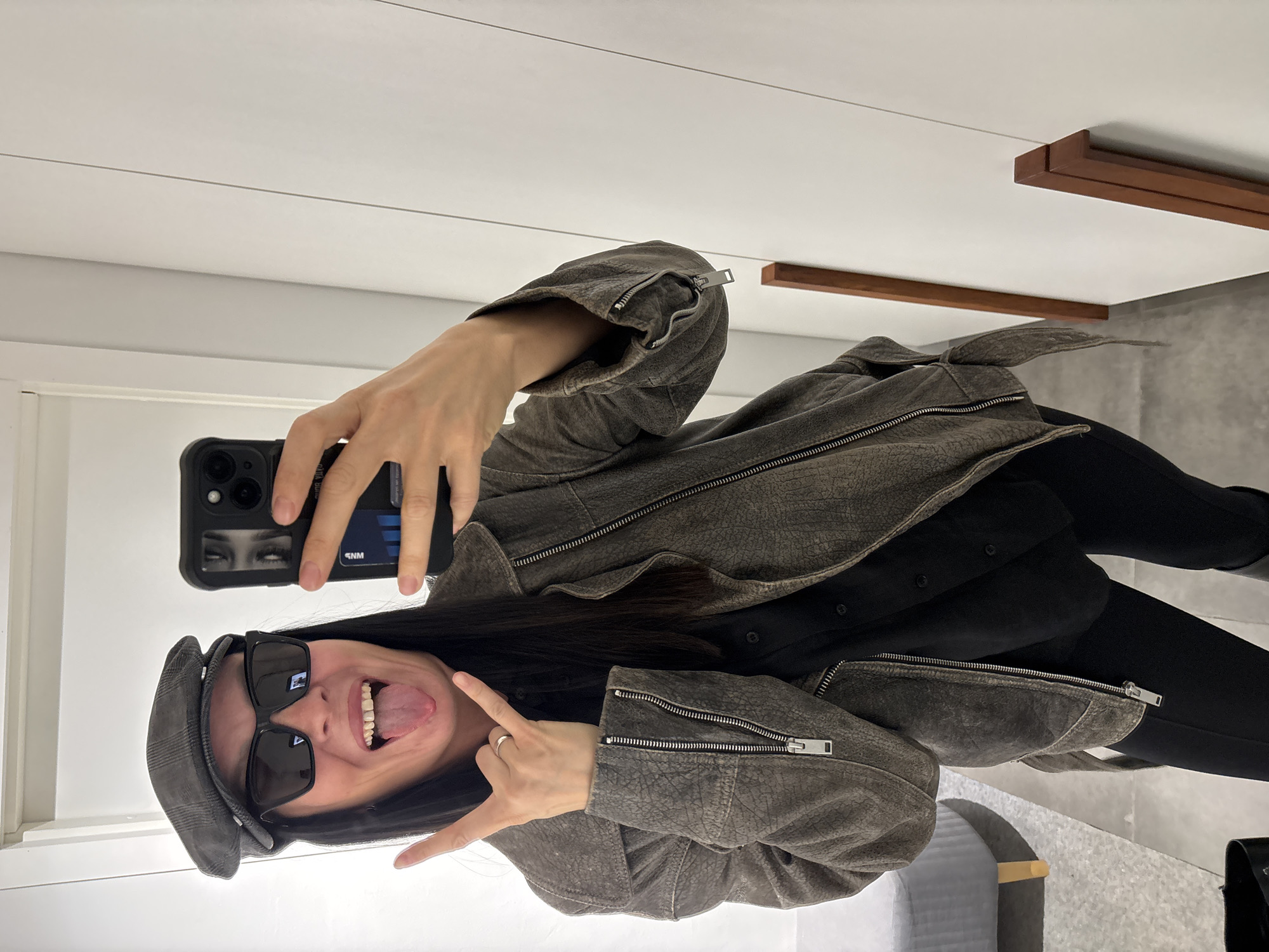 A person takes a selfie in a room. The person is wearing sunglasses, a cap, and a leather jacket. They are sticking their tongue out and making a hand gesture. The room has white walls and a light-colored floor. There is a wooden accent on the ceiling. A gray upholstered seat is visible. A credit card is visible in the person's phone case. The image does not provide information about the city of Chaikovsky, Russia.