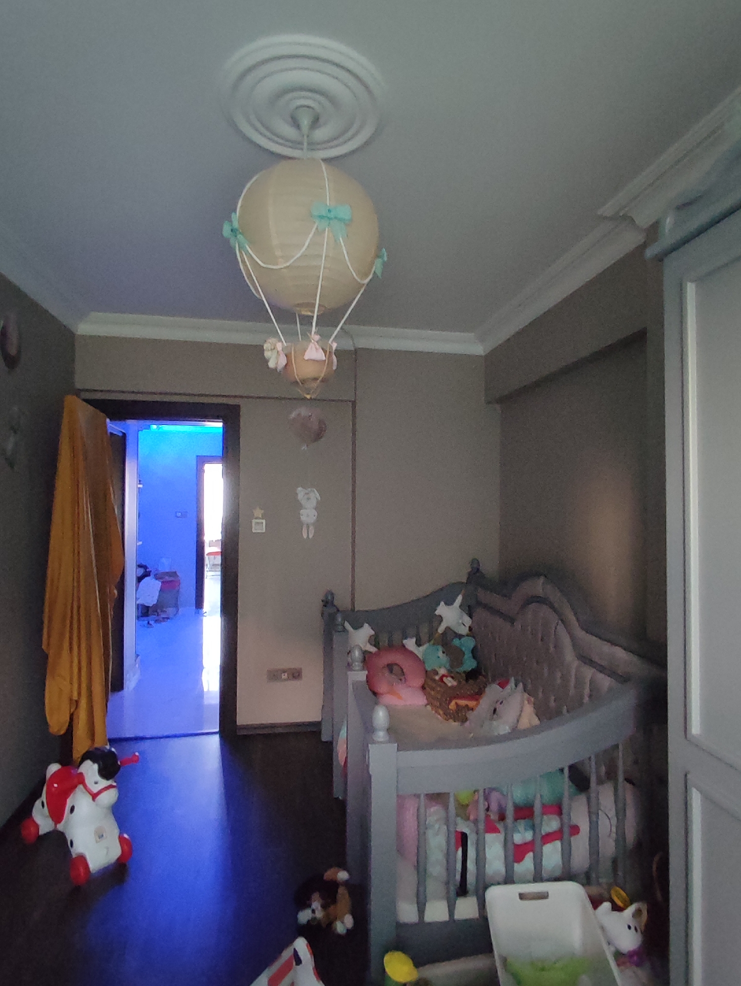 This image depicts an indoor scene, likely a child's room, located in Kartal, Turkey. The room is furnished with a crib filled with stuffed animals and bedding. A rocking horse toy is situated on the floor in the foreground. A doorway on the left side of the image leads to another room illuminated with bright blue light. A decorative hot air balloon-shaped lamp hangs from the ceiling. The walls are painted a neutral color, and the floor appears to be dark wood. The overall impression is of a well-lit, playful environment designed for a child.