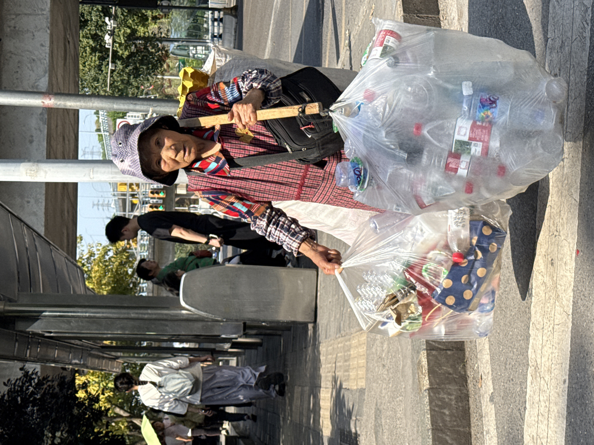 An elderly woman, likely a waste collector, is captured mid-stride on a sunny day in Nan'an District, China. She is the central figure, looking directly at the viewer with a weathered but composed expression. She wears a checkered long-sleeved shirt, a blue and white checkered bucket hat, and a black shoulder bag.
Her main activity involves carrying two large, clear plastic bags filled with recyclables, predominantly plastic bottles and other refuse. These bags are suspended from a sturdy wooden stick, which she holds horizontally across her body with both hands, using it as a carrying pole.
The setting is an urban street or sidewalk. In the background, other people are visible, some sitting on what appear to be public benches or at a bus stop. There are trees, street poles, and glimpses of buildings and traffic lights, indicating a bustling city environment. The bright sunlight casts strong shadows on the ground, suggesting it's daytime.
Notable details include the practical, homemade carrying system for the recyclables, highlighting a common practice in resource recovery. Inside the bags, various labels and packaging are visible on the plastic bottles and other items; while specific text is mostly unreadable due to resolution and being inside bags, some labels feature red, blue with yellow polka dots, and other colorful designs, suggesting diverse beverage and product containers. The overall scene conveys a slice of daily life and labor in a Chinese urban setting.