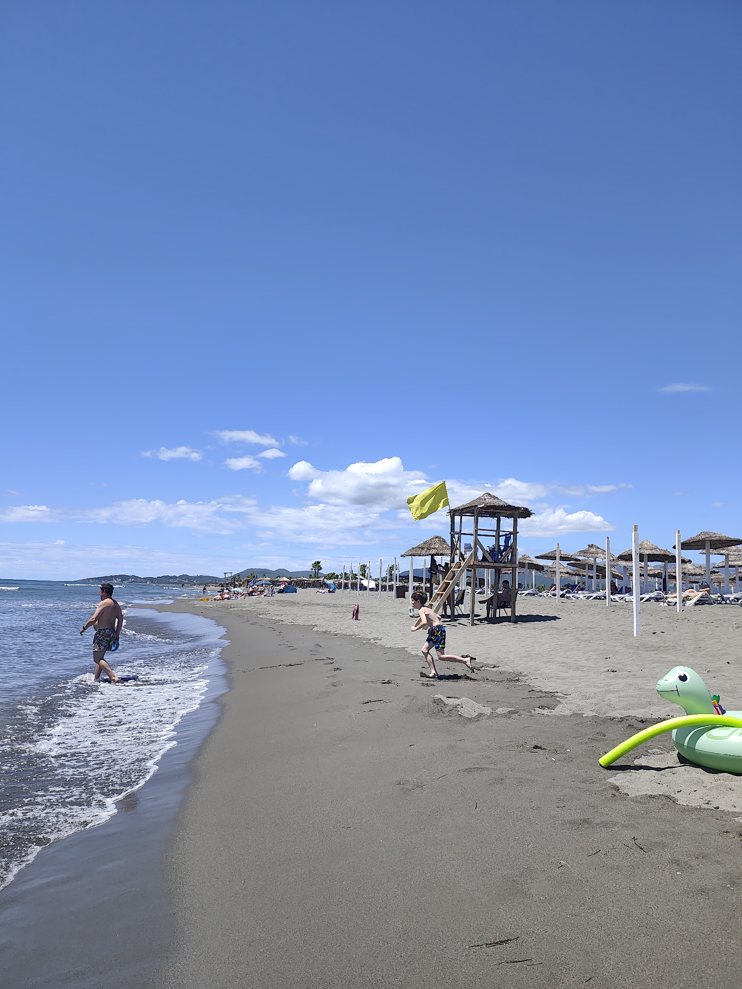 A sunny day at a beach in Donji Štoj - Shtoji i Poshtëm, Montenegro, with a clear blue sky and scattered white clouds. The sandy beach has dark grey sand and is lined with rows of thatched-roof umbrellas and lounge chairs. In the foreground, a man walks in the shallow water along the shoreline. Further up the beach, a young boy runs towards a wooden lifeguard tower with a thatched roof. A yellow flag flies from the top of the tower. In the background, other beachgoers are visible, some sitting under umbrellas and others walking along the water's edge. A green inflatable turtle toy with a yellow ring is on the sand to the right of the frame. The sea is calm with small waves.