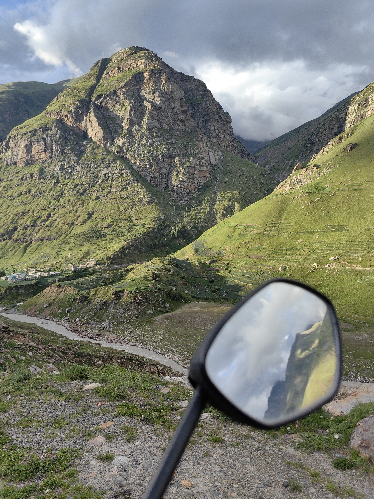 A motorbike's side mirror reflects a dramatic mountain landscape in Yari-Khoksar, India. The mirror shows a jagged, sun-drenched mountain peak with green slopes and a cloudy sky. In the foreground of the main image, the mirror is attached to a black handle, positioned over a rocky, gravelly terrain dotted with sparse green vegetation and small purple flowers. Beyond the mirror, a winding, light-colored river flows through a valley. The valley walls are steep and covered in lush green grass, with evidence of terraced farming on the slopes. A small cluster of buildings, likely a village, is visible in the mid-ground on the left side of the valley. The sky above is a mix of dark, brooding clouds and patches of bright sunlight, casting shadows across the mountains. The overall scene depicts a remote and rugged natural environment.