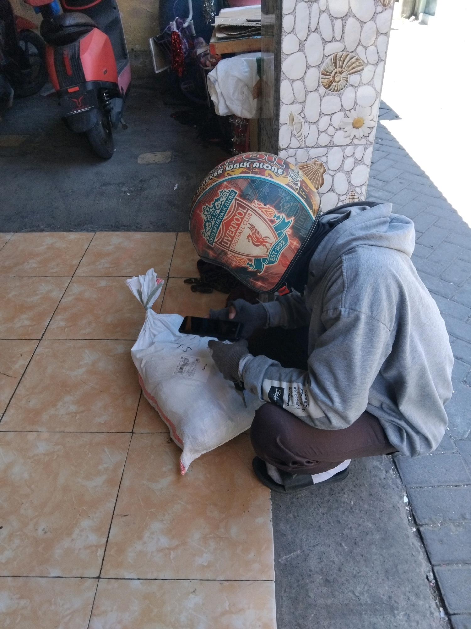 In Kepuh Kiriman, Indonesia, a person wearing a gray hoodie and brown pants crouches beside a white bag on a tiled floor. The person also wears a helmet adorned with the Liverpool Football Club logo and the slogan "You'll Never Walk Alone." The background shows a red scooter, a wall decorated with shells and a flower, and a paved area. The person appears to be interacting with a smartphone, which is placed on top of the white bag.