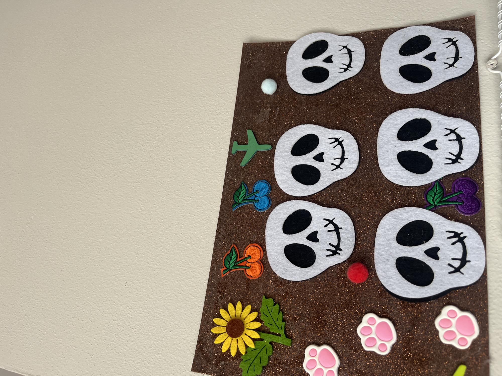 This is a close-up, eye-level, indoor shot of a brown glitter-covered board with various felt cutouts adhered to it. The board is attached to a beige wall. The cutouts include a yellow sunflower with green leaves, two orange cherries with green leaves, two blue cherries with green leaves, a green airplane, a small light blue pompom, a small red pompom, three white paw prints with pink centers (two visible in full, one partially visible), and seven white skull cutouts with black eyes and mouths. The skulls are arranged in two rows, with four in the top row and three in the bottom row. The board appears to be part of a larger display or craft project. There is no indication of the city of Onokivtsi, Ukraine, or any forensic details beyond the described objects.