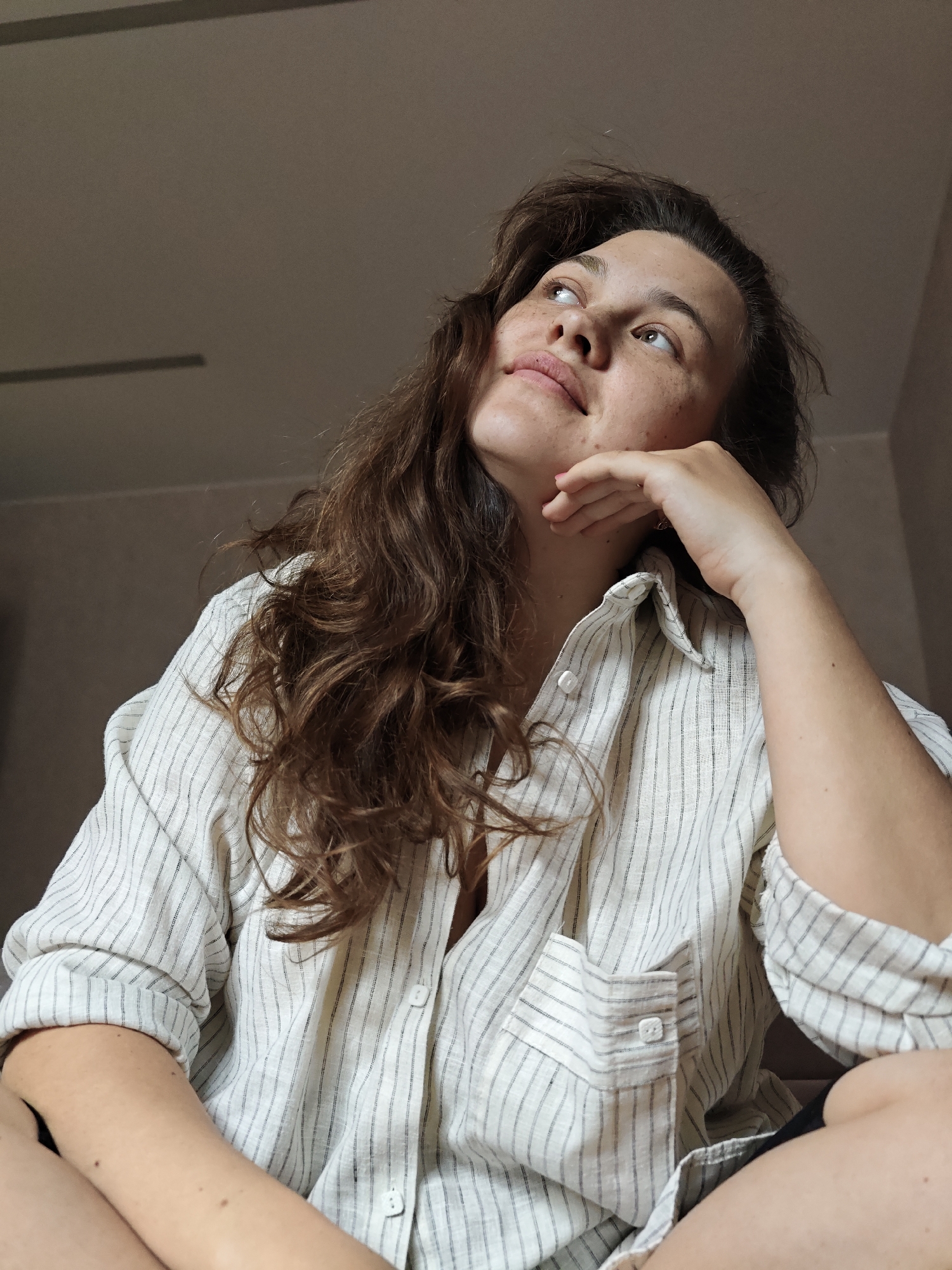A young woman with long, wavy brown hair and freckles is seated, leaning on her right hand as if in thought. She is wearing a light-colored, vertically striped button-down shirt. The room appears to be indoors, with a plain, light-colored wall or ceiling in the background. Her gaze is directed upwards. Given the prompt's request to include the location, the scene is set in Chaikovsky, Russia, though this is based on the prompt's information and is not visually verifiable from the image alone.